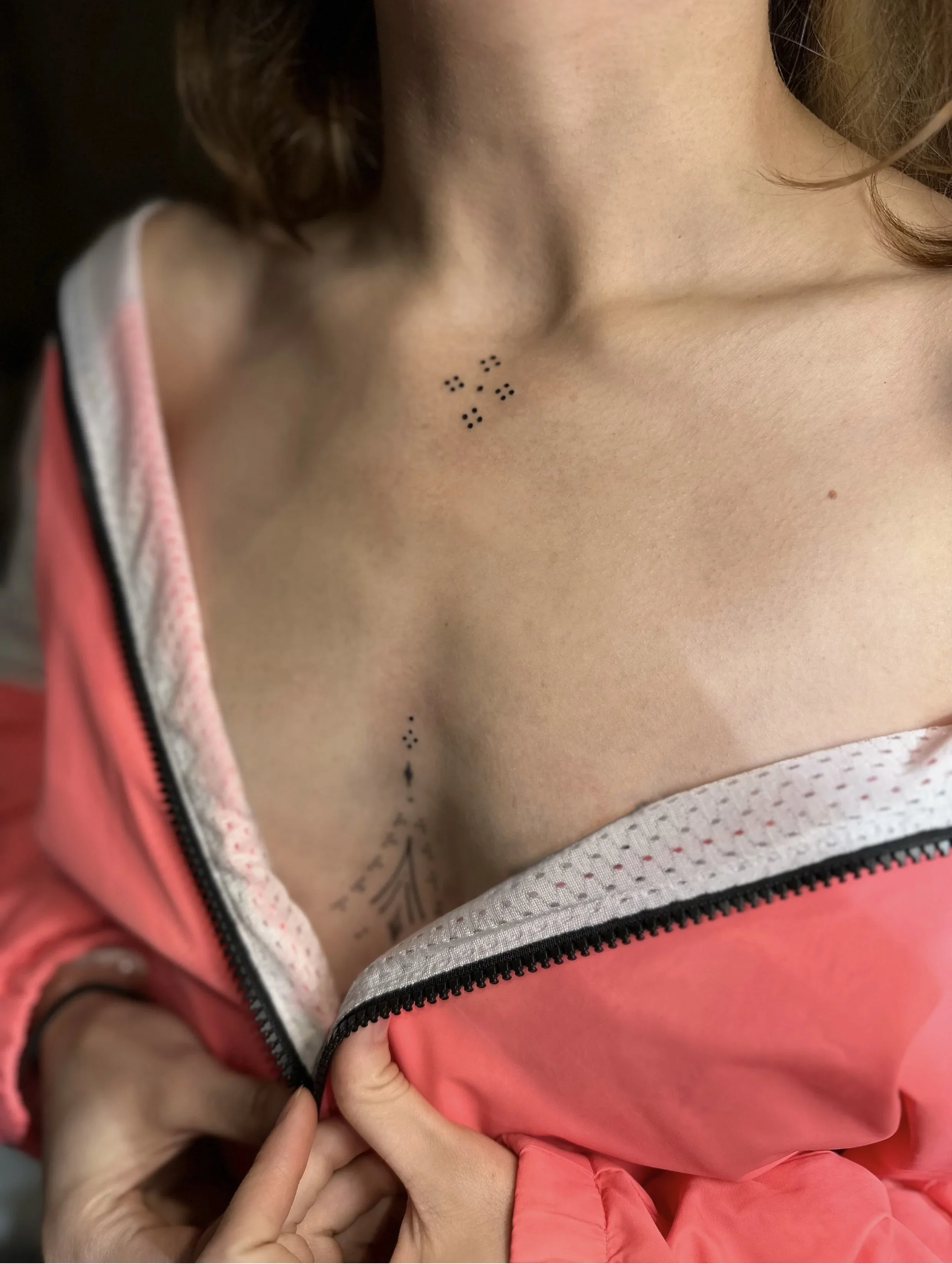 Close-up of a person's upper chest revealing small braille dots tattooed on the skin, partially covered by a pink jacket and a hand resting nearby.