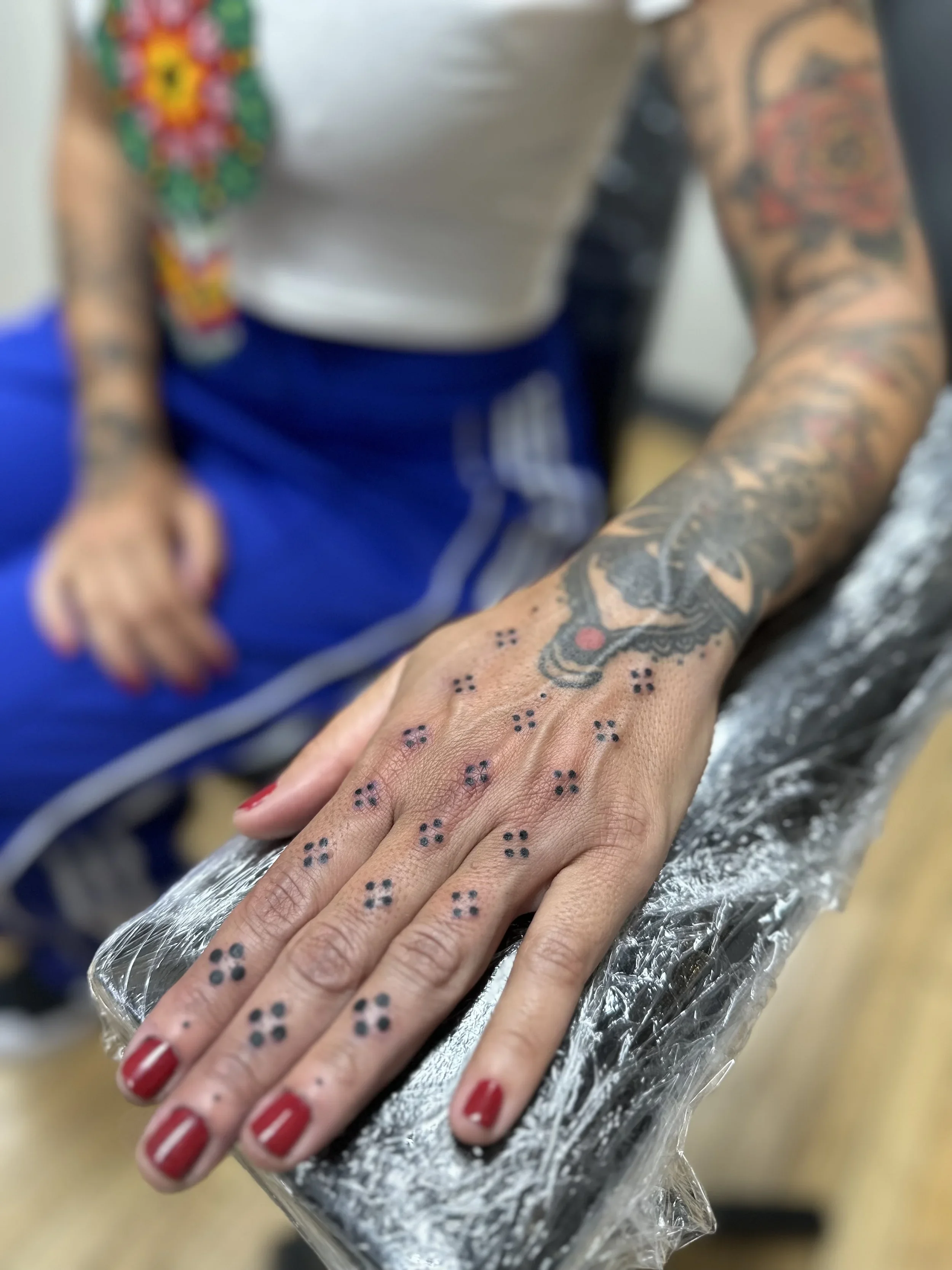Close-up of a person's hand with a tattoo and red nail polish, resting on a surface covered with aluminum foil. The background shows part of a person's arm with tattoos and a blurred shirt.