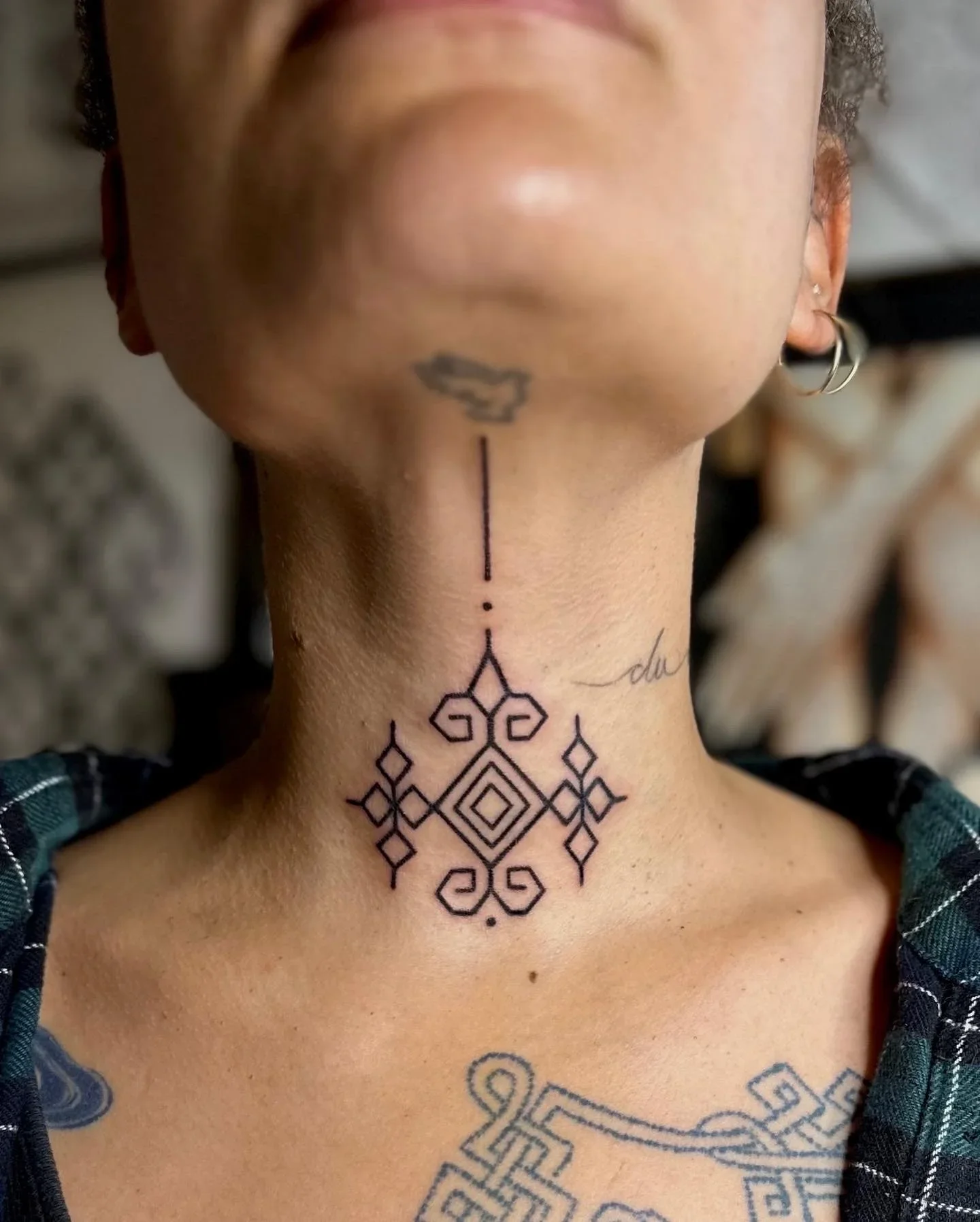 A person's neck with multiple tattoos including geometric patterns, a small cloud, and some script, along with earrings and a plaid shirt collar visible.