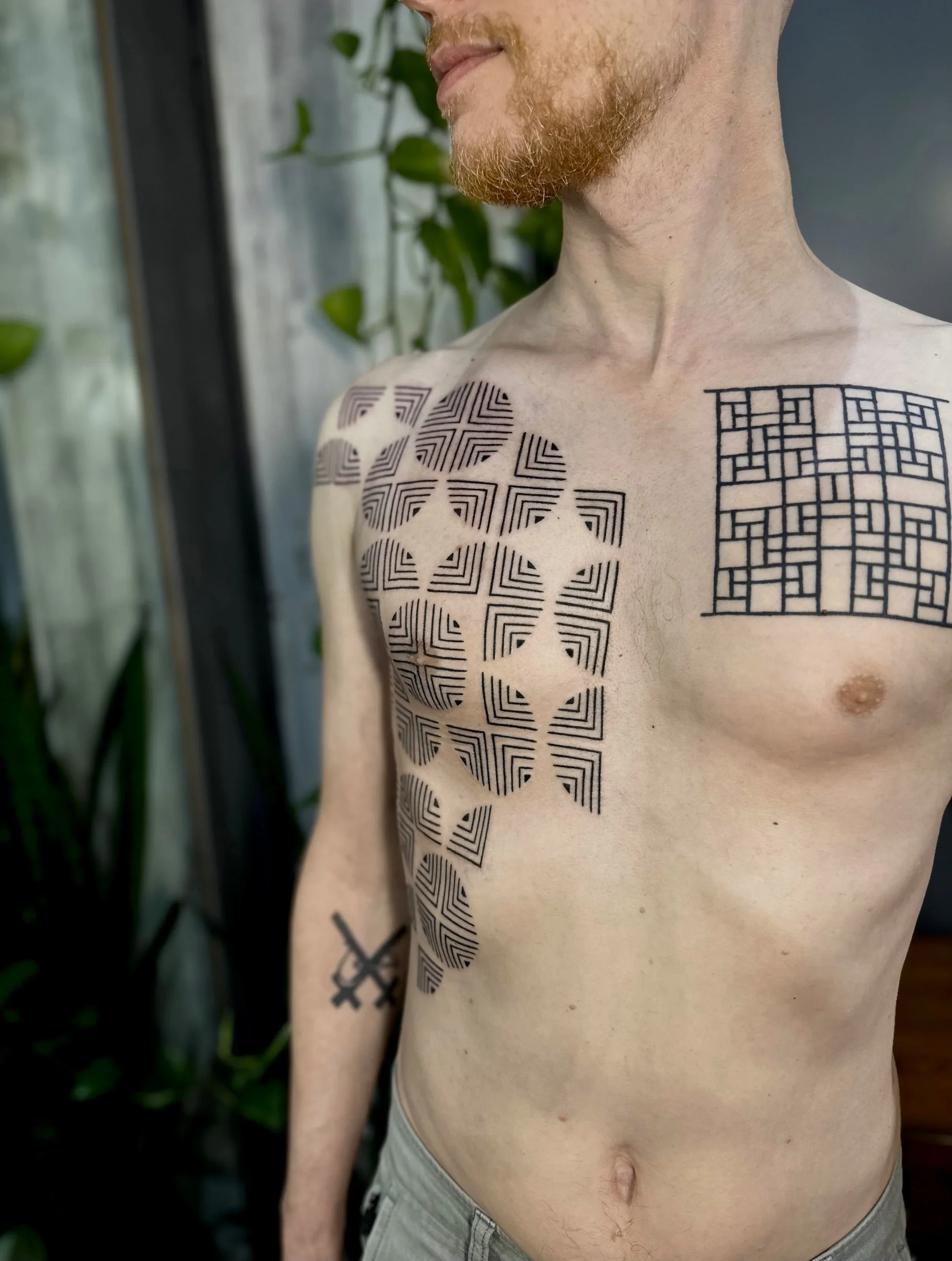 A shirtless man with red hair and tattoos on his chest and arm standing indoors near a wall with green plants in the background.