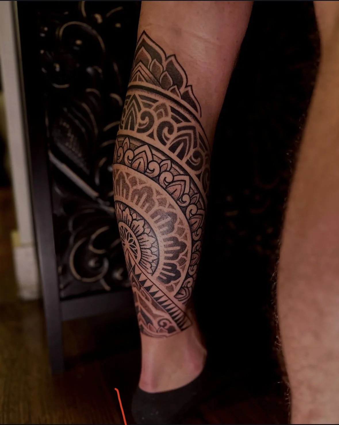 A person's leg with an intricate black and white geometric tattoo, featuring mandala and floral patterns, standing on a wooden floor with a black ornate metal gate in the background.