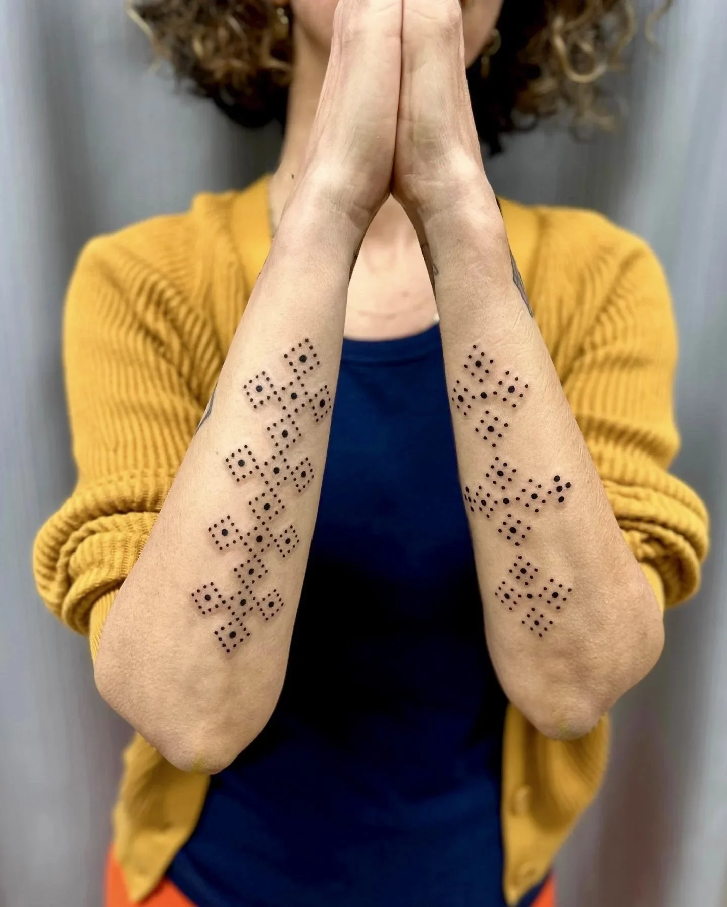 Person with tattoos on forearms, palms pressed together, wearing a yellow cardigan and dark top, with curly hair, against a plain background.