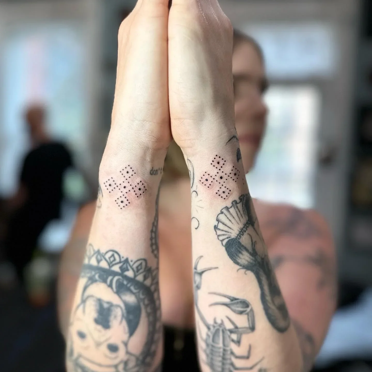 Person with tattoos holding palms together in front of face, tattoos include dotted designs, skull, and other images, with a blurred background.