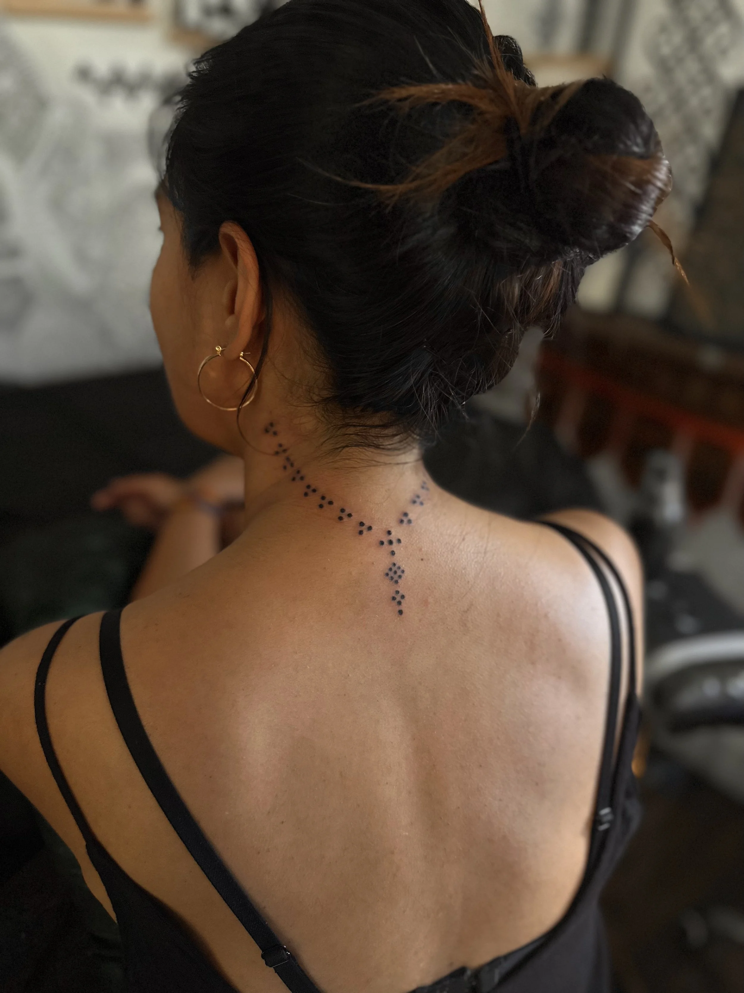 Rear view of a woman with dark hair tied up, wearing black spaghetti strap top, earrings, and a dotted tattoo on her neck and shoulder.