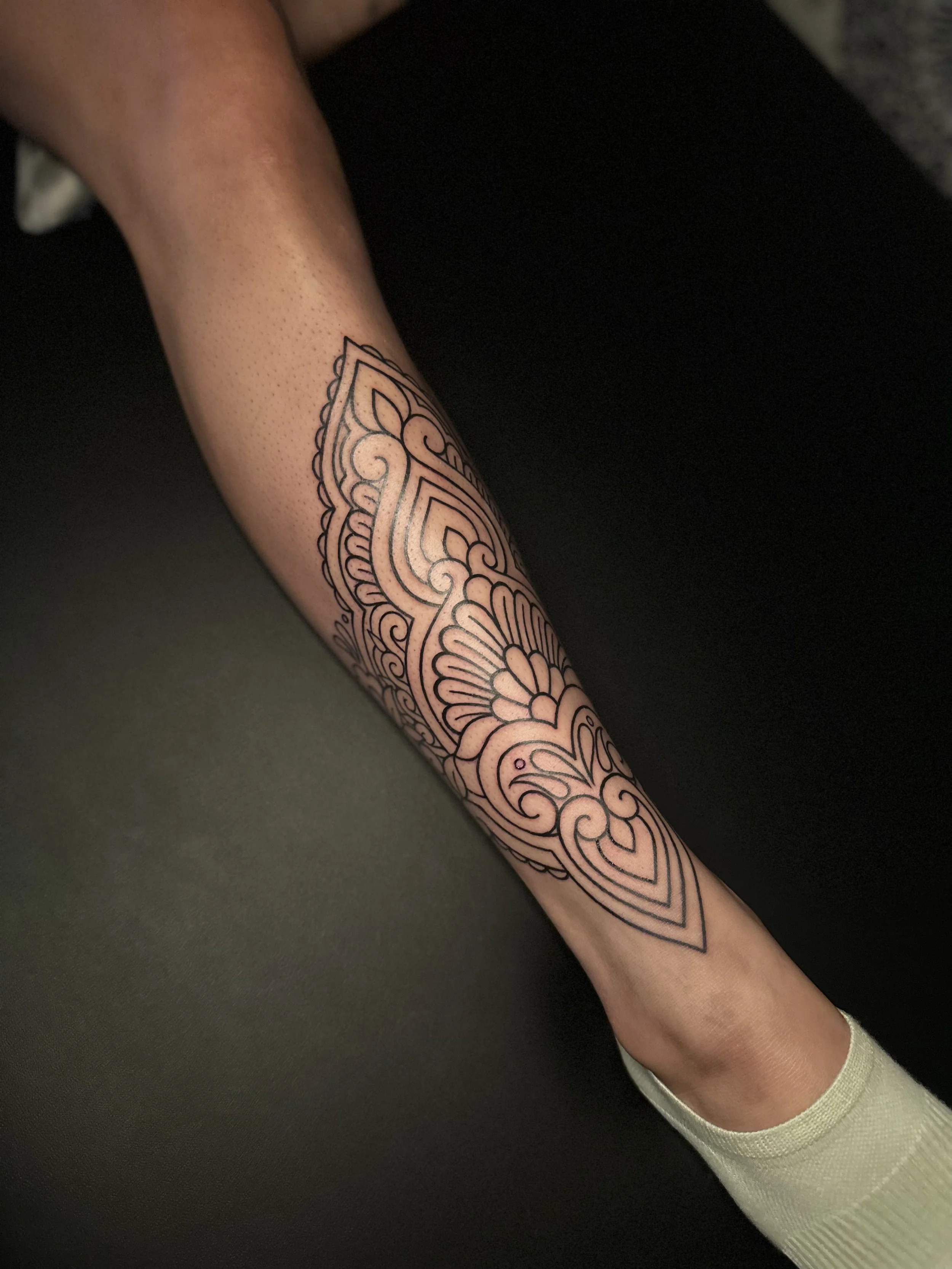 A person's arm with a detailed black and white henna tattoo sleeve, resting on a dark surface. The person is wearing a light-colored long-sleeve shirt.