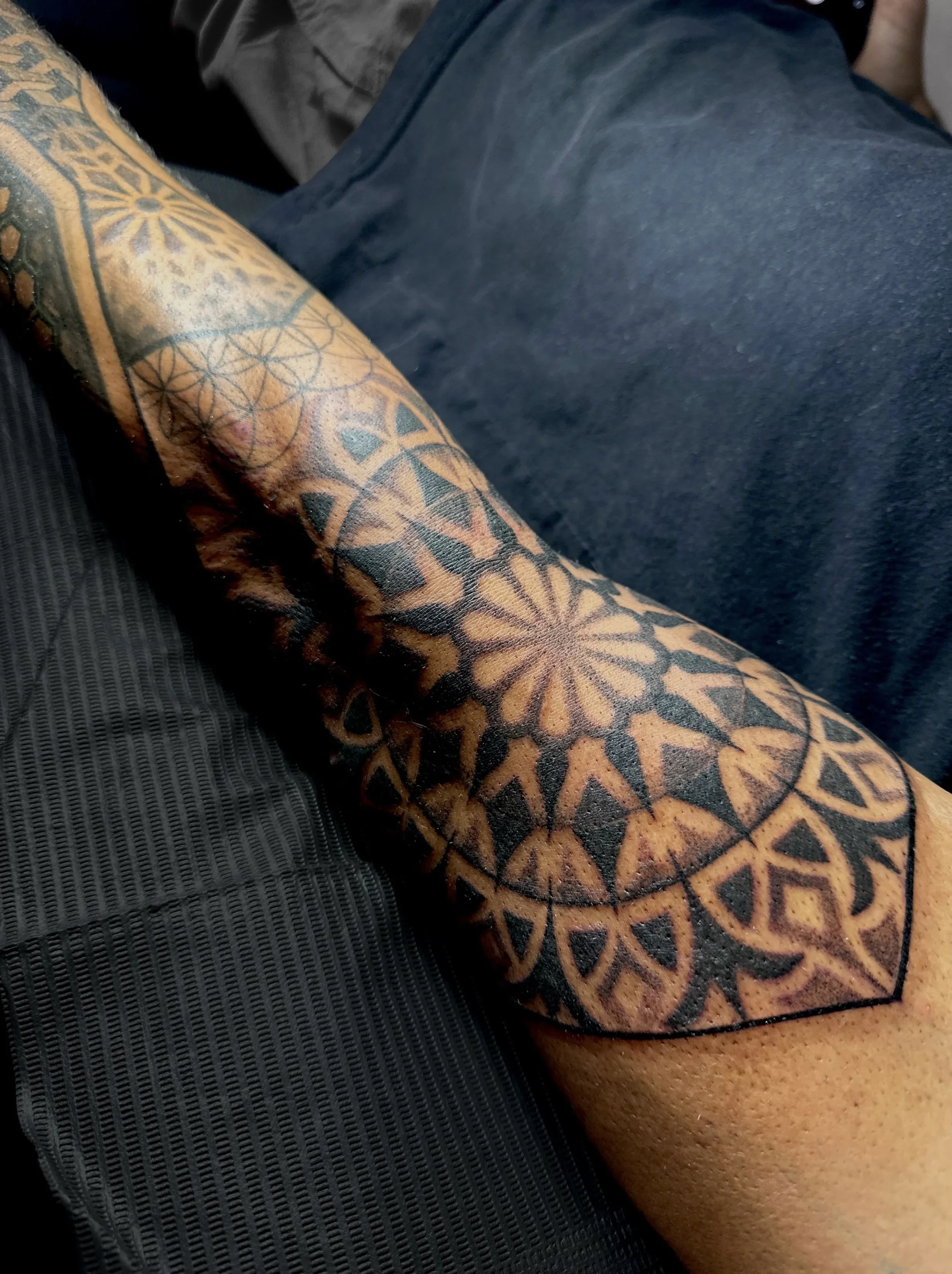 Close-up of a tattooed arm with geometric and floral designs, resting on a textured dark surface.