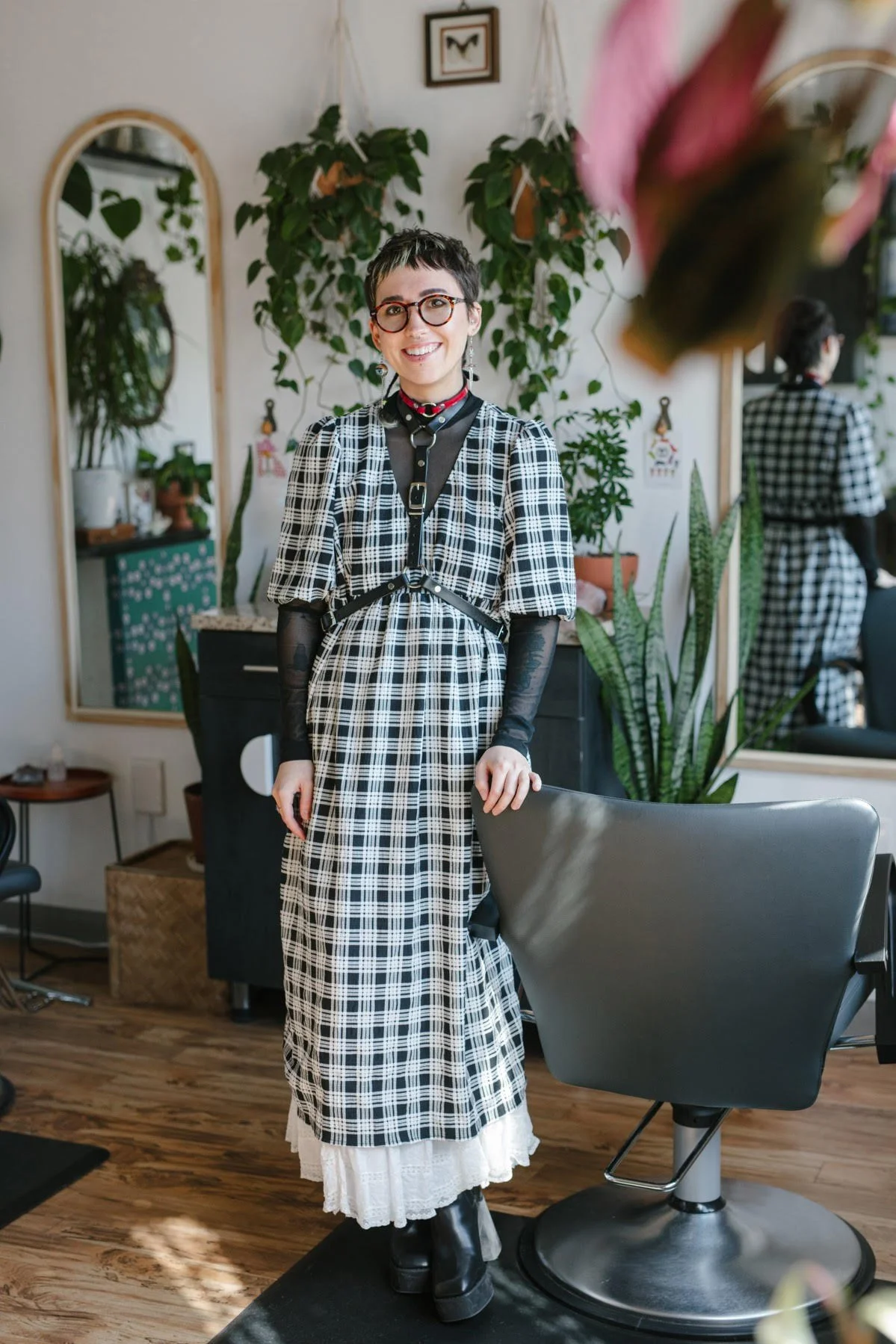 A young woman with short dark hair, glasses, and a smile, standing in a hair salon. She wears a black and white plaid dress layered over a white lace skirt, with black boots and a harness-style accessory. The salon has plants, mirrors, and a wooden floor.