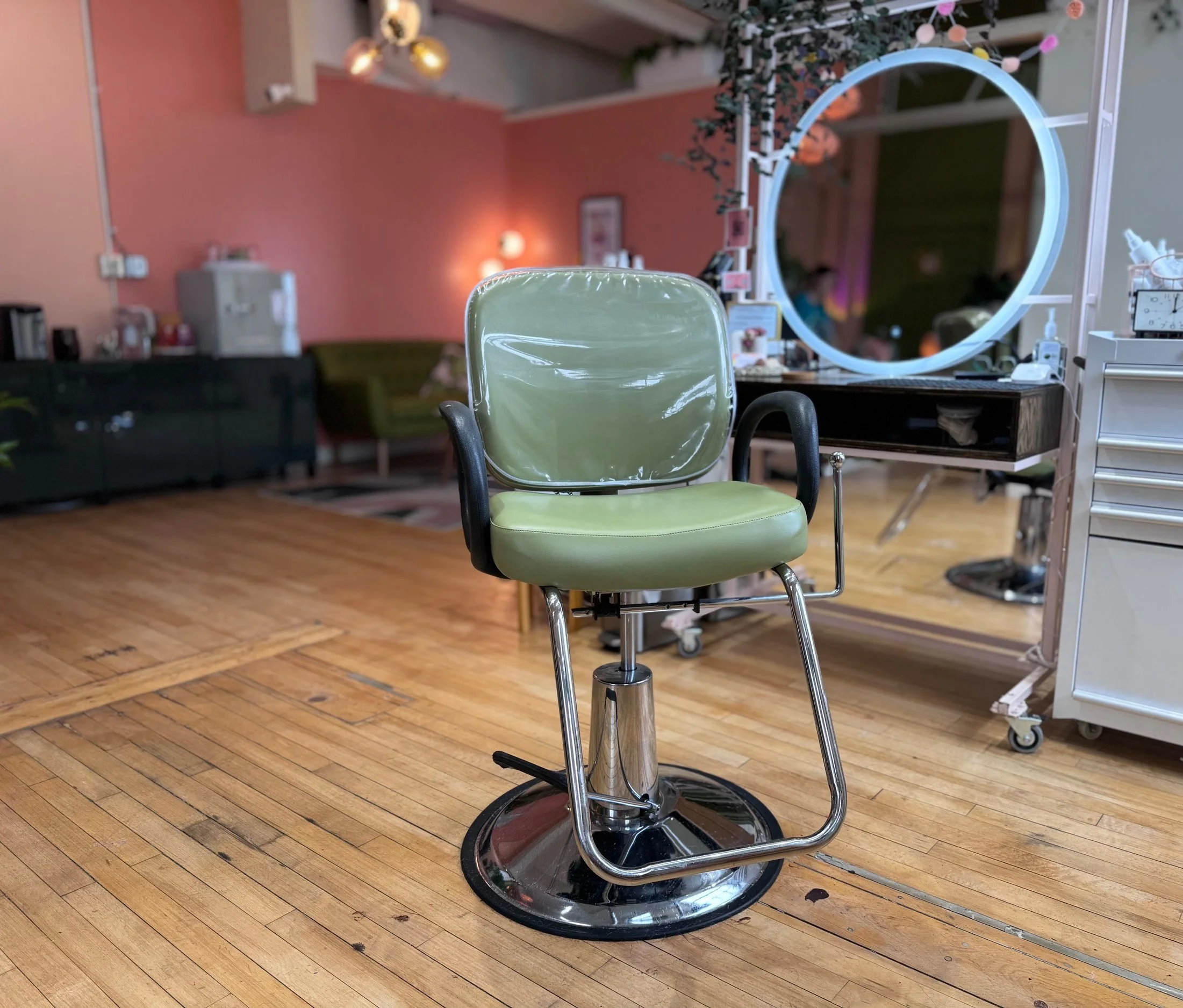 Empty green barber or salon chair in a modern salon with wooden floors, a large round mirror, and pink walls in the background.