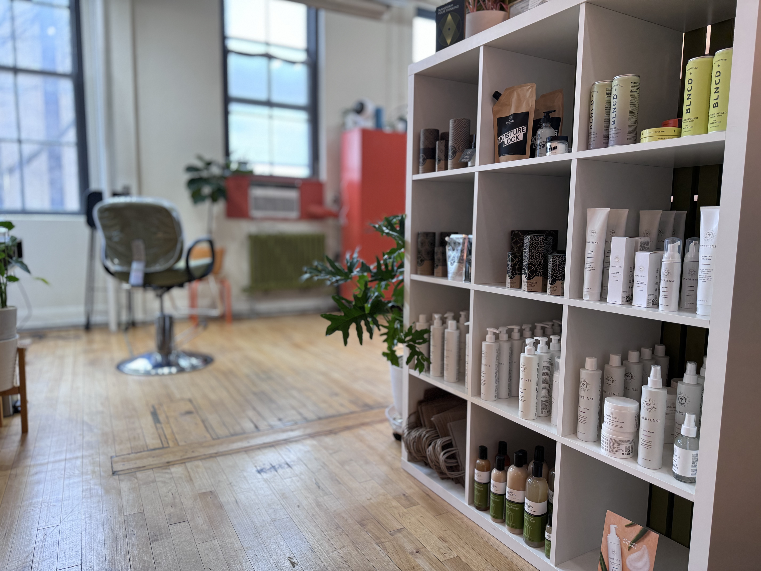 Interior of a wellness or beauty salon with a white display shelf containing skincare and haircare products, a vintage wooden chair, a potted plant, large windows, and a red radiator on the wall.