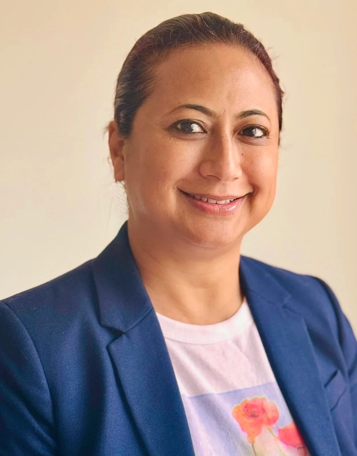 A woman with dark brown hair pulled back, wearing a dark blue blazer over a white t-shirt with a floral print, smiling against a plain beige background.