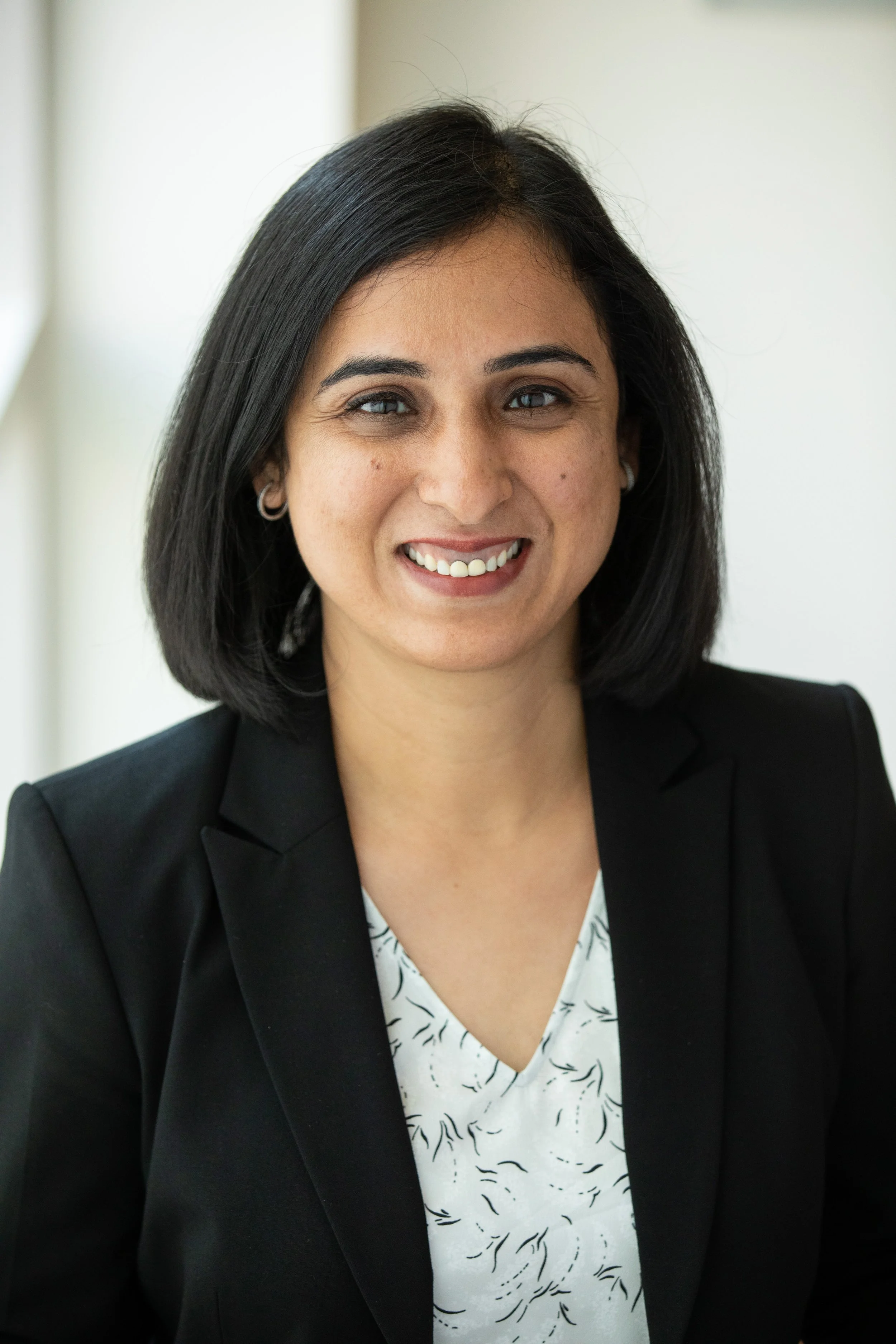 A woman with black hair, wearing a black blazer and a white patterned blouse, smiling at the camera.