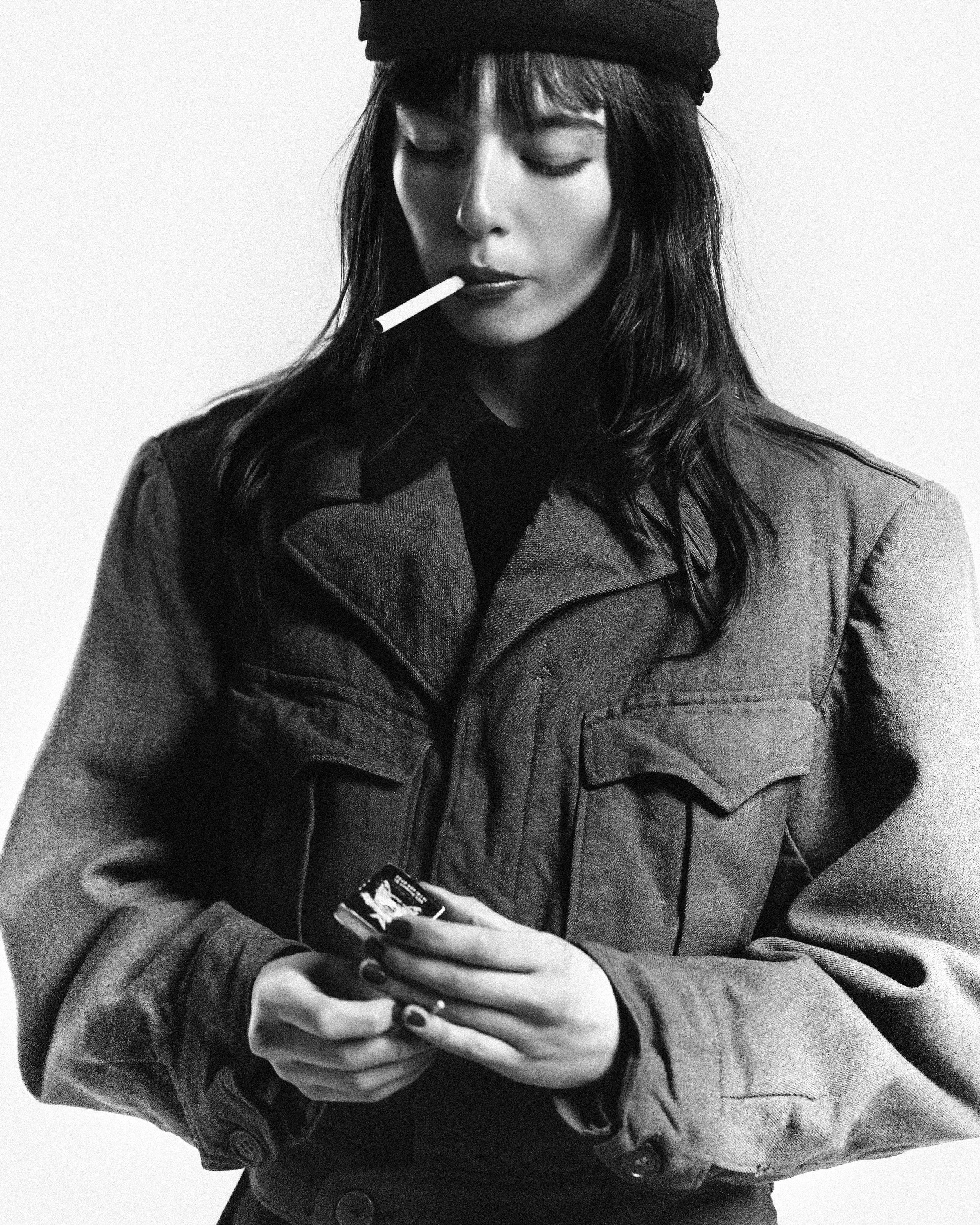 Black and white photo of a woman wearing a beanie and jacket, holding a cellphone in one hand and a cigarette in her mouth.