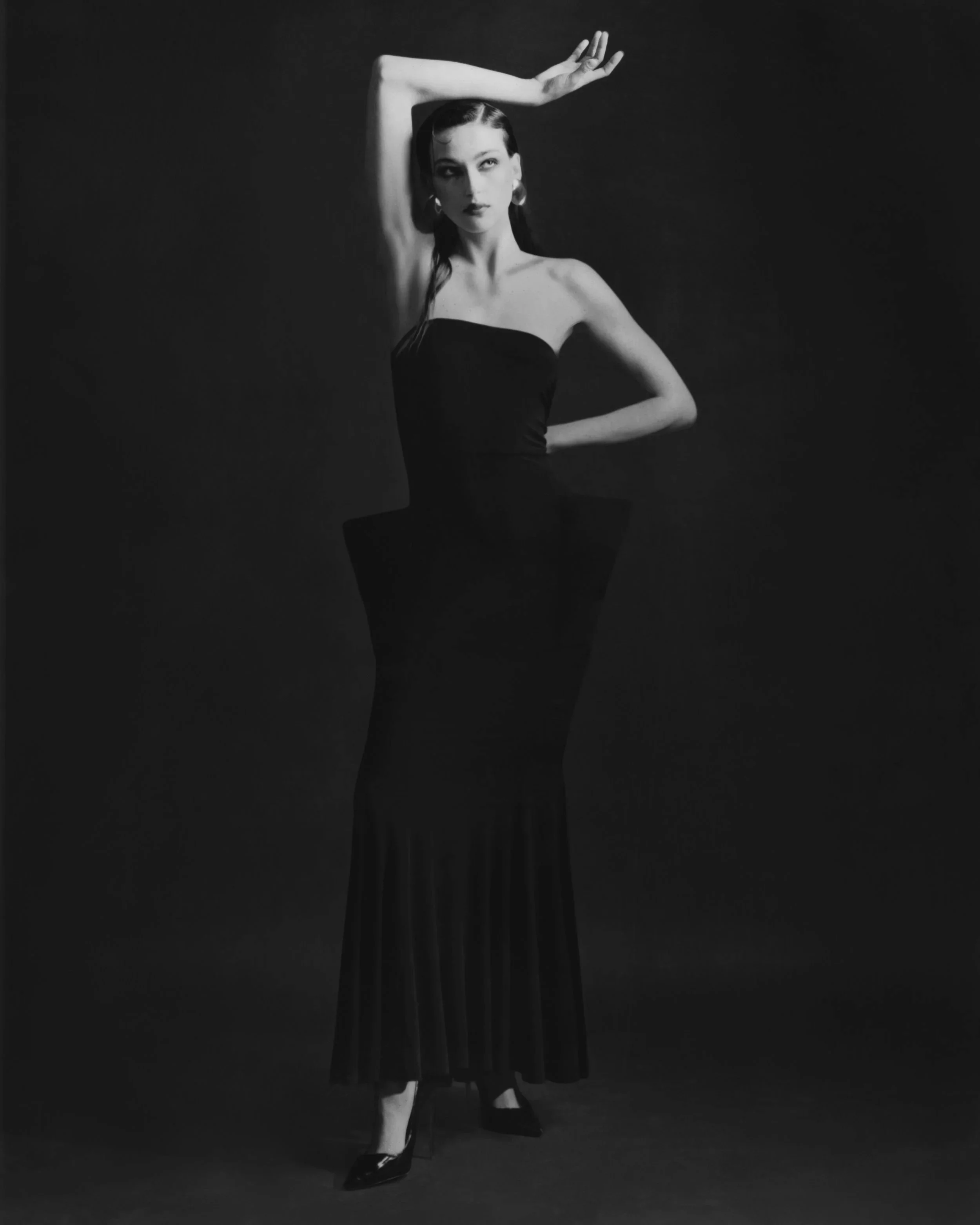 Black and white photo of a woman in a strapless dress with one arm raised above her head, posing with a confident expression against a dark background.