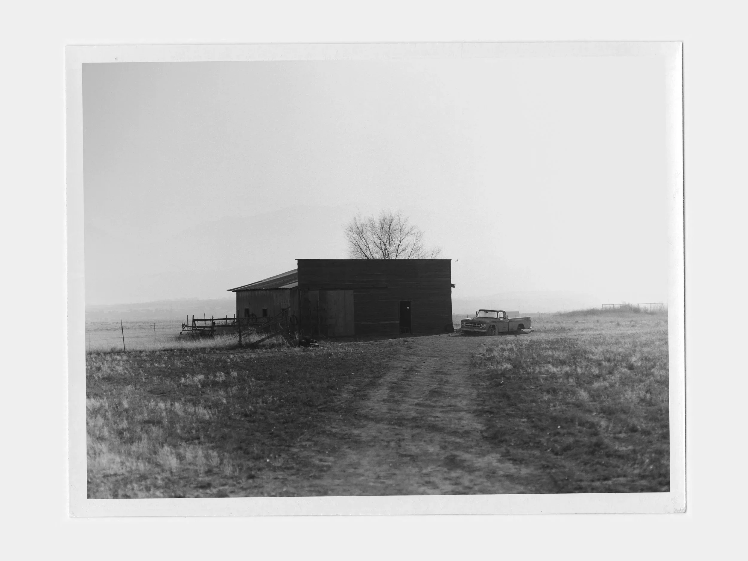 A black and white photo of a rural landscape with a dirt road leading to an old wooden barn, a vintage truck parked nearby, and a leafless tree in the background, under a mostly clear sky.