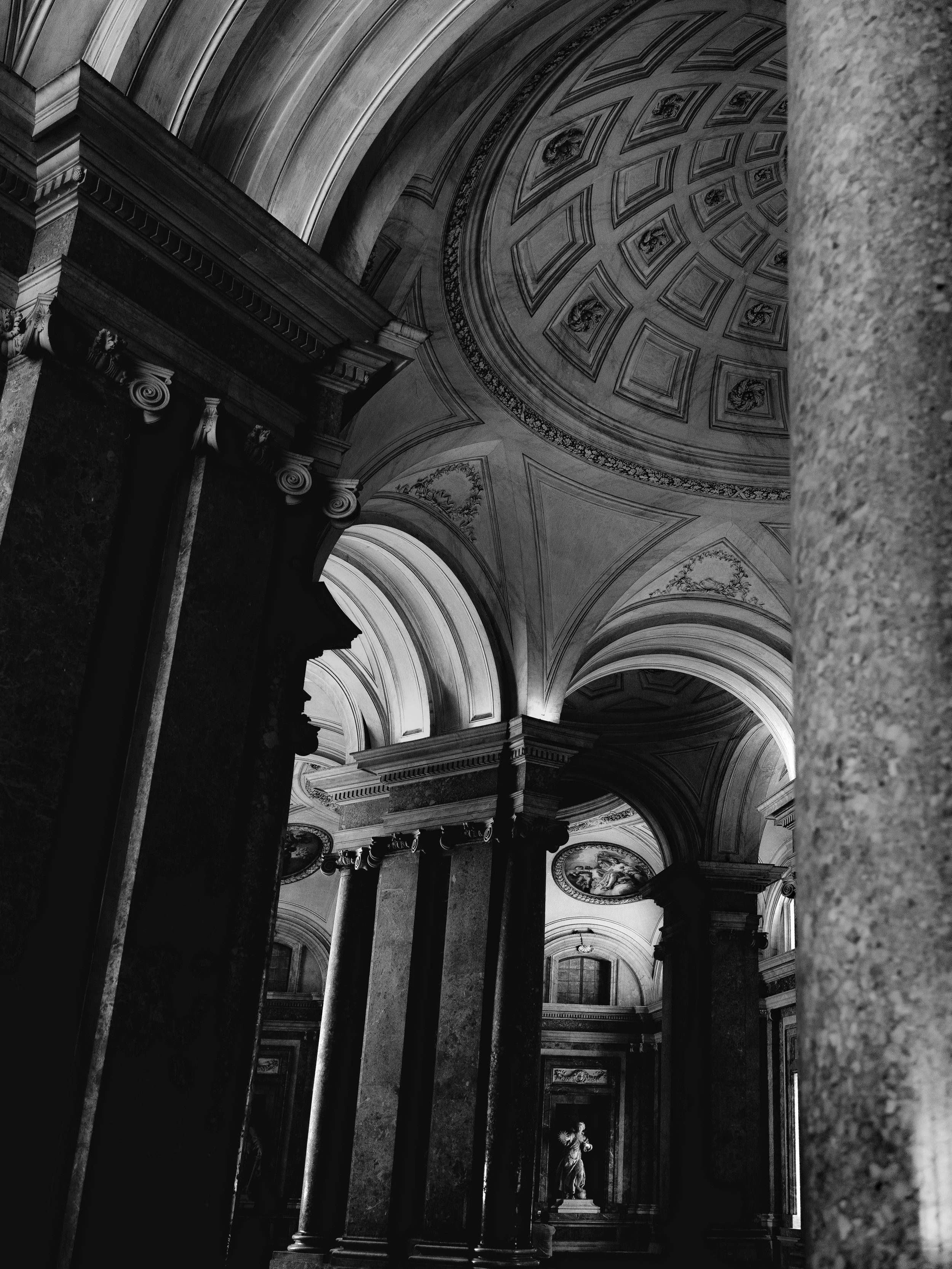 Interior view of a grand, classical-style building with tall columns, arches, and ornate ceiling detail in black and white.