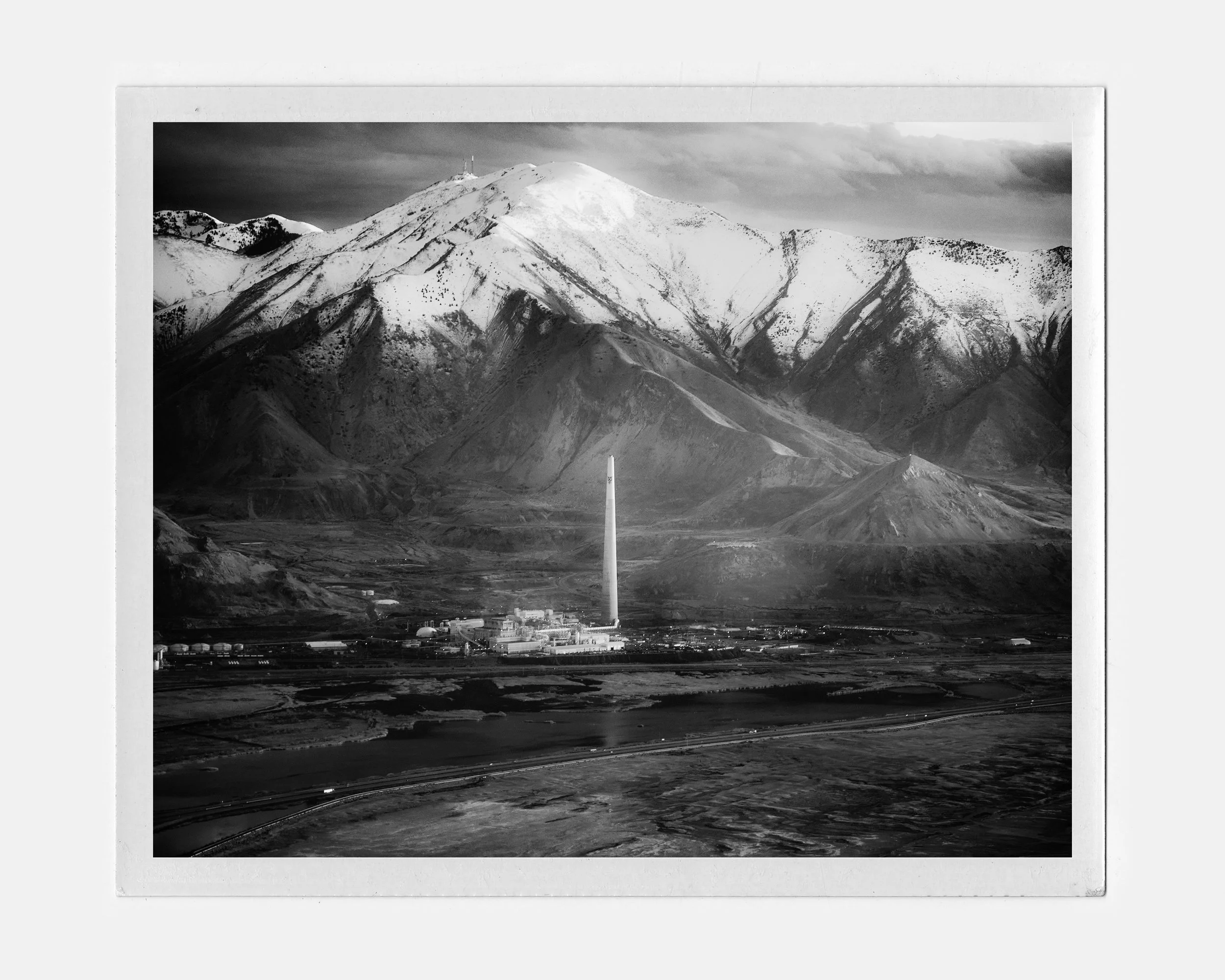 Black and white photo of a mountainous landscape with snow-capped peaks and a power plant with a tall chimney in the foreground.