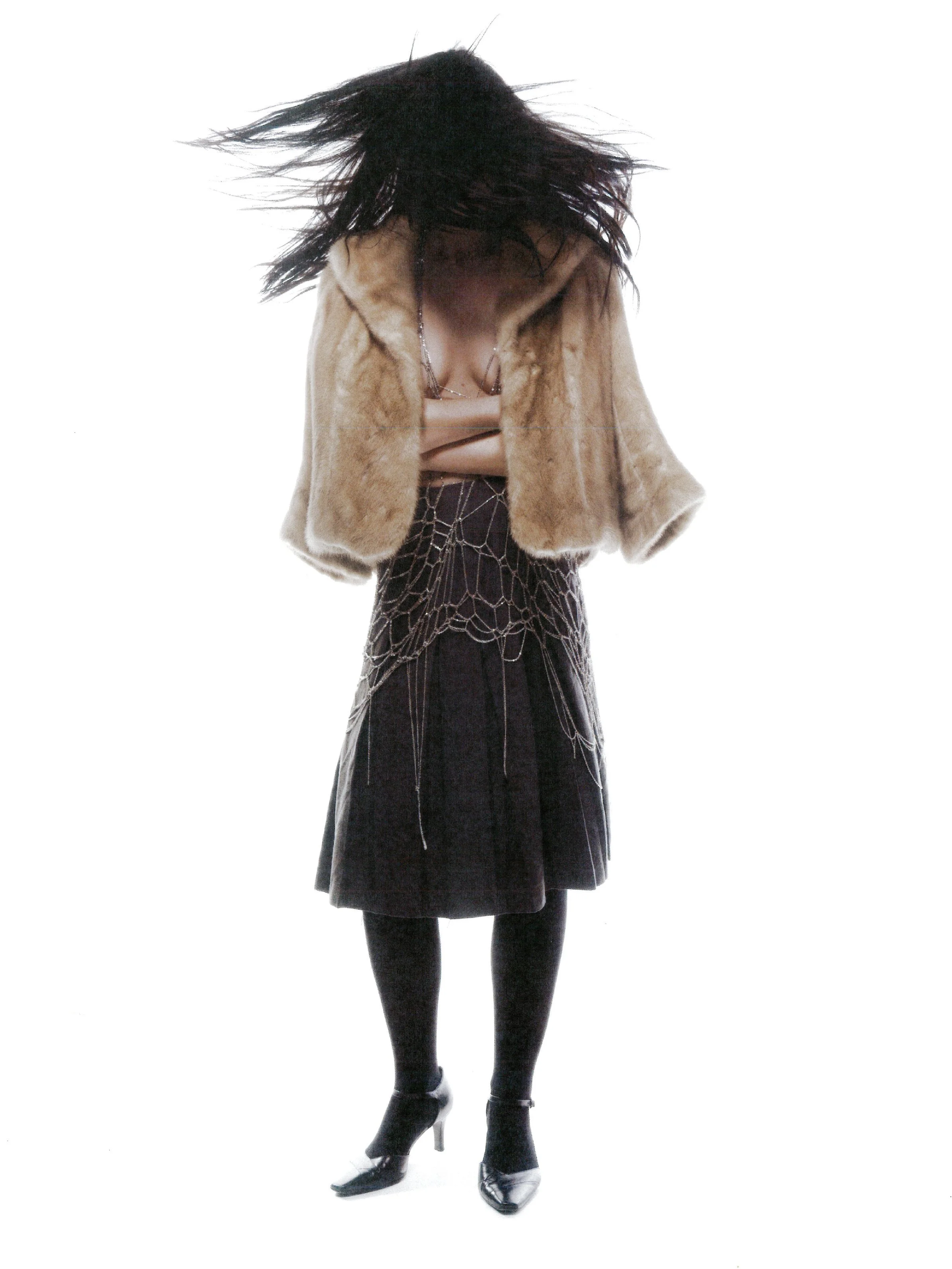 A woman with long dark hair is flipping her hair forward, obscuring her face. She is wearing a beige faux fur coat, a beige top, a black pleated skirt with a spiderweb pattern, black tights, and black high heels.