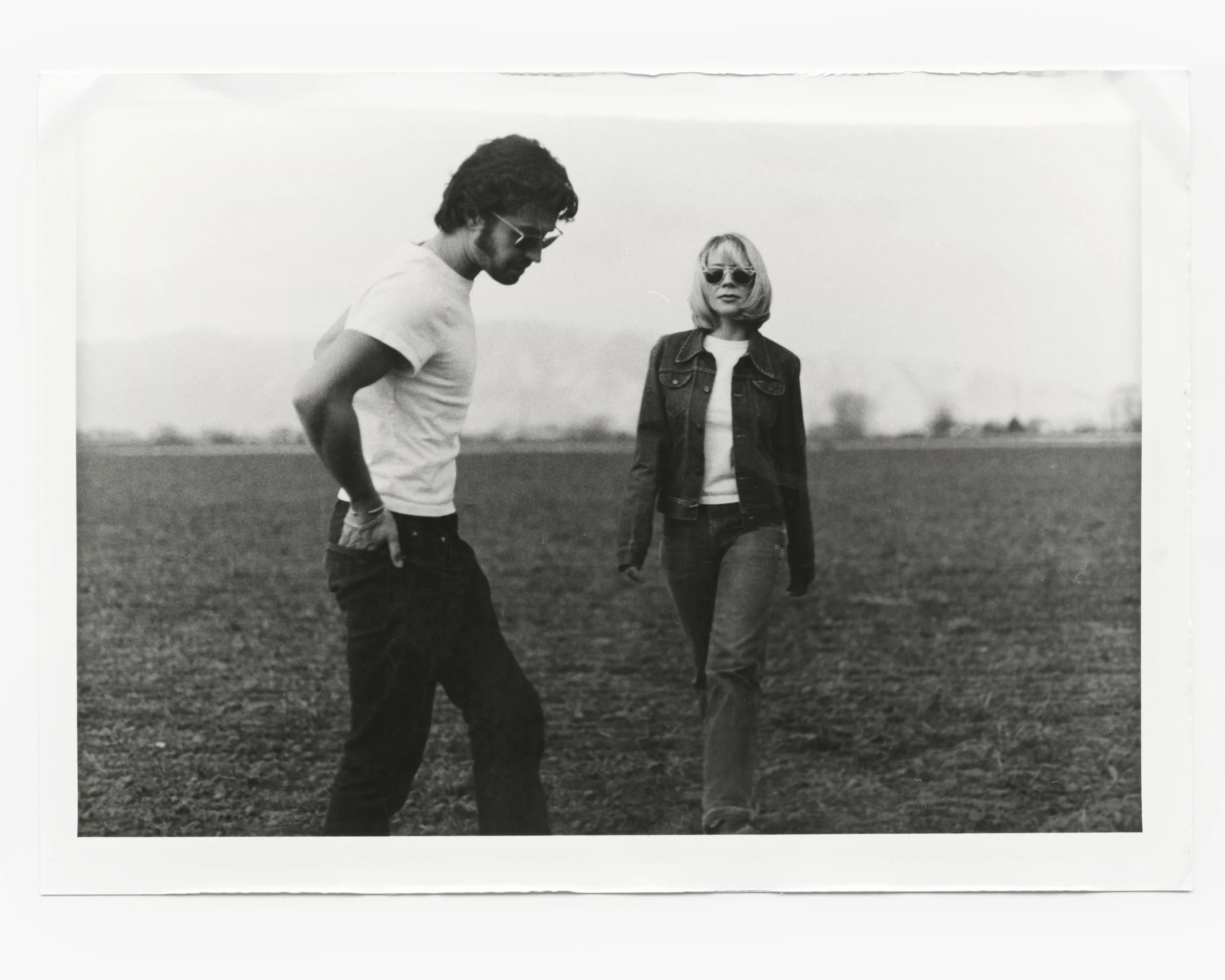Black and white photo of a man and woman walking in an open field on a cloudy day. The man is looking down with hands in pockets, wearing sunglasses, a t-shirt, and dark jeans. The woman is walking toward the camera, wearing sunglasses, a denim jacke