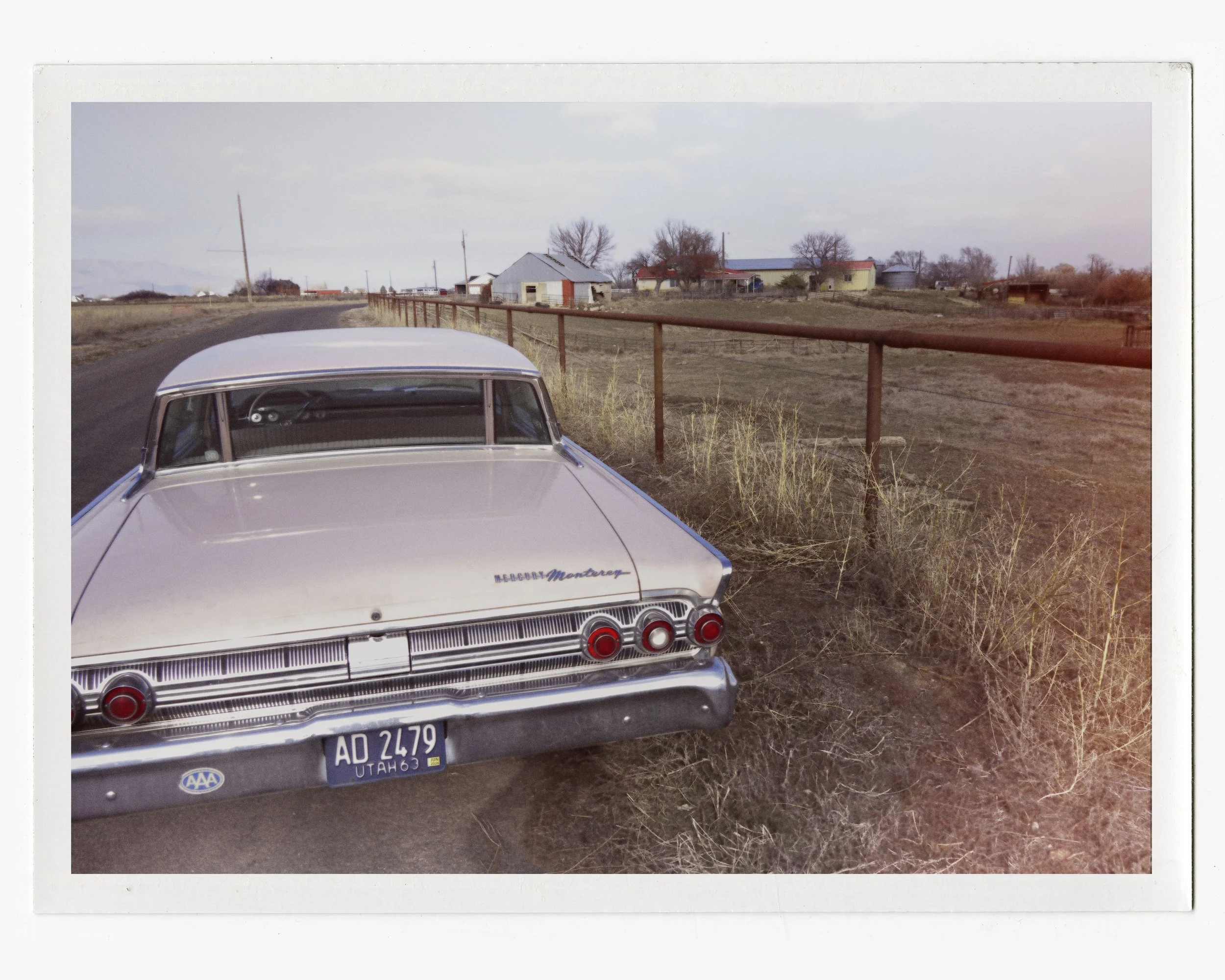 Vintage white Mercury Monterey parked on side of rural road with dry grass and a red railing, with farms and leafless trees in background.