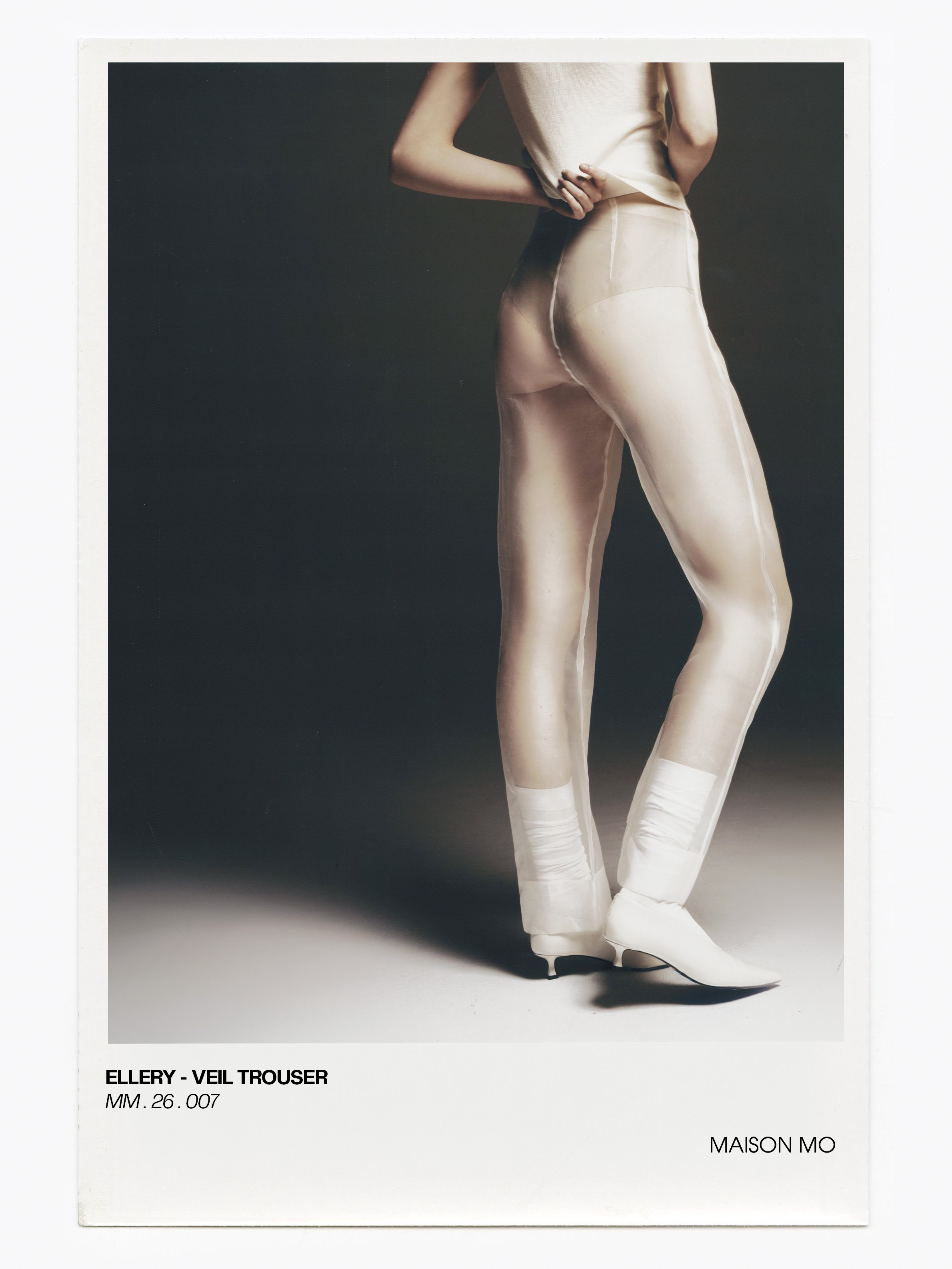 A person wearing sheer veil trousers and white shoes, holding their top with one hand behind their back, against a dark background.