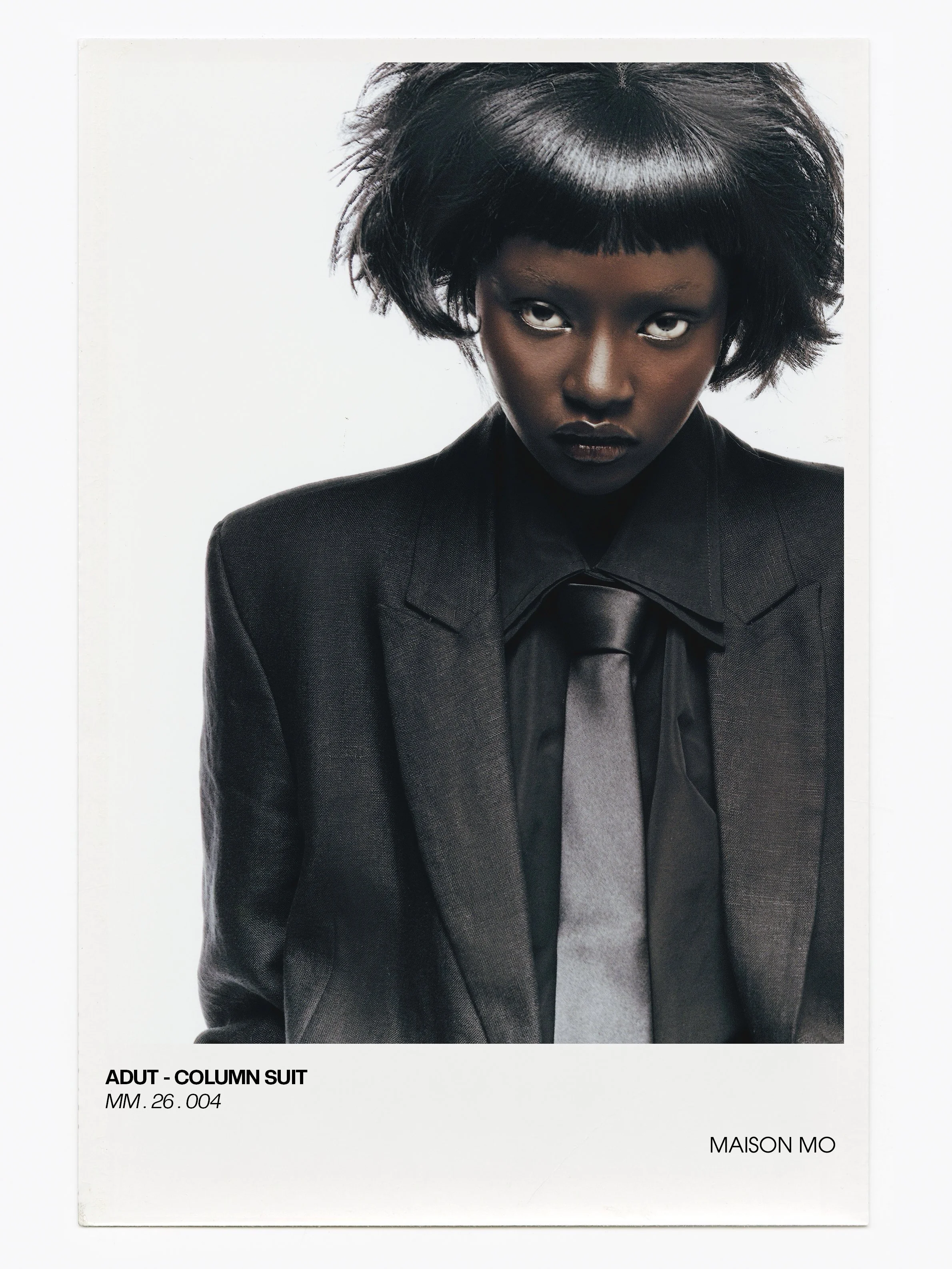 A woman with dark skin wearing a black suit, black shirt, and black tie, looking directly at the camera with a serious expression against a plain white background.