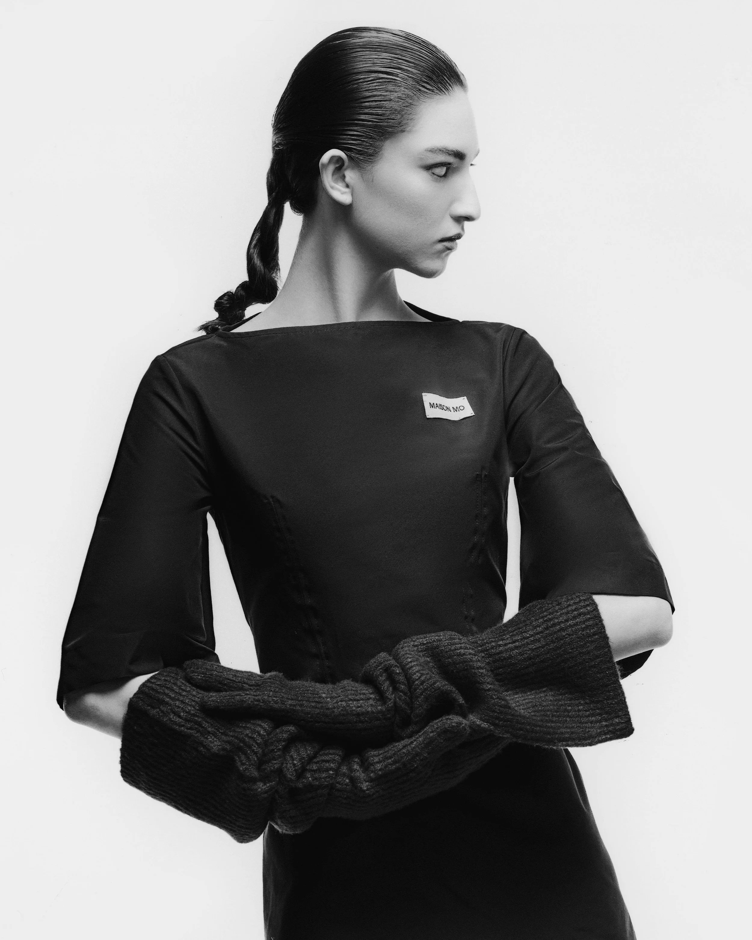 Black and white photo of a woman with slicked-back hair in a braid, wearing a long-sleeved black top with sleeves pulled down over her hands, standing against a plain background.