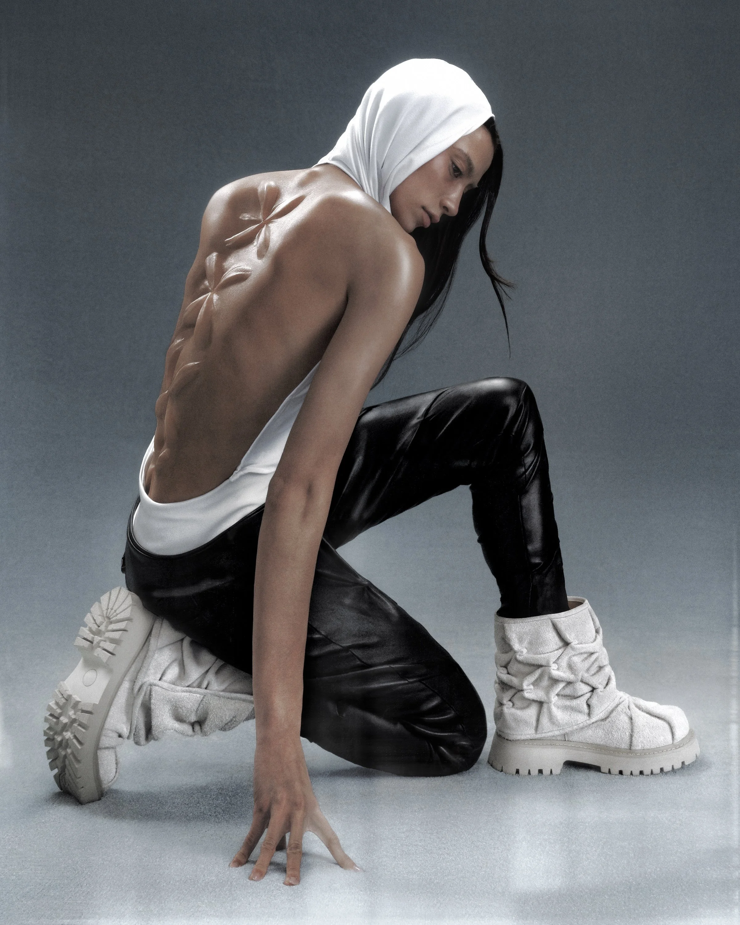 A woman wearing a white headscarf, black leather pants, and white sneakers, kneeling on the floor, looking down with her back and abs exposed.