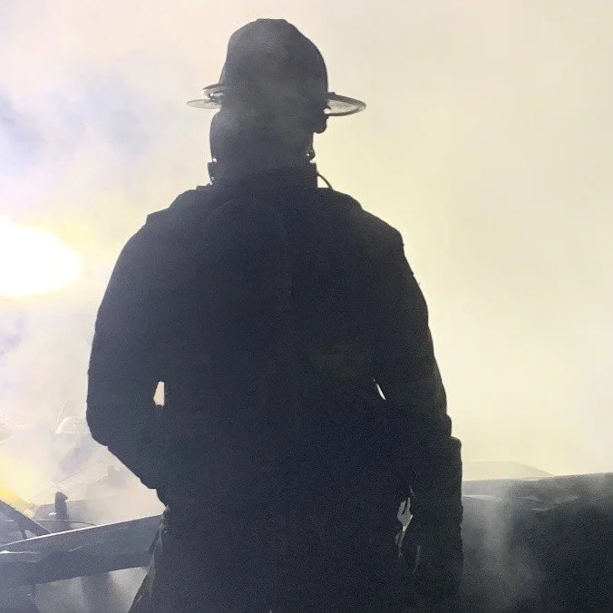 A firefighter paramedic wearing a cap with a circular brim, standing in a smoky environment with bright light behind them, facing away from the camera.