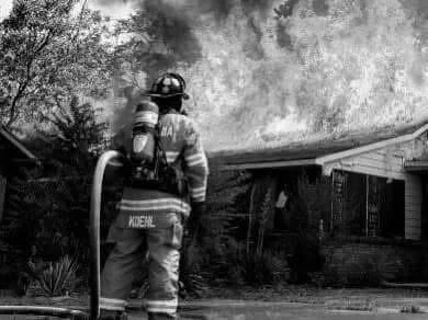 Firefighter battling a house fire, with flames and smoke coming from the building.