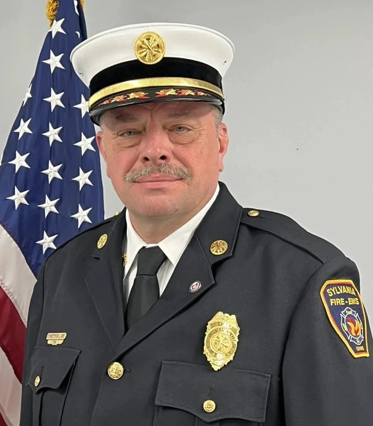 A firefighter in formal dress uniform standing in front of an American flag, with a patch on the uniform indicating association with Sylvania Fire-EMS.
