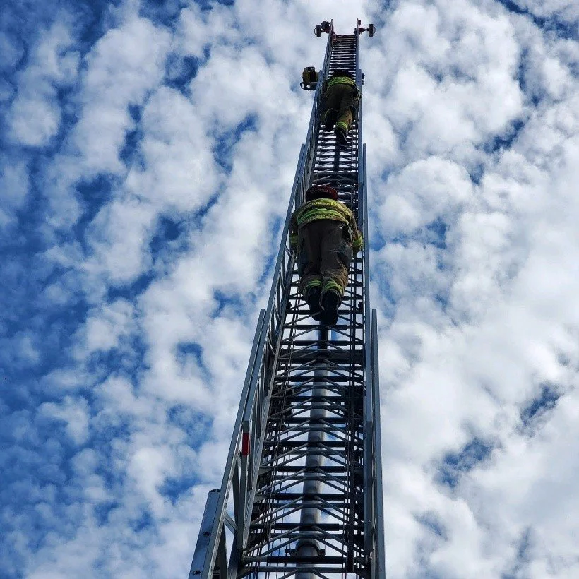 Two firefighters climbing a tall extension ladder against a cloudy sky.