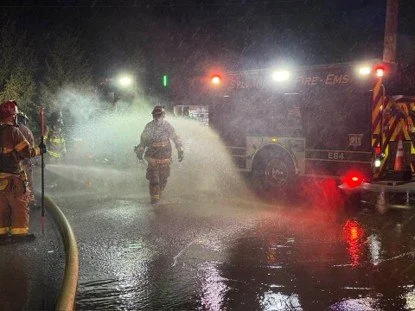 Firefighters working at night with water on the street, fire truck with flashing lights in the background.