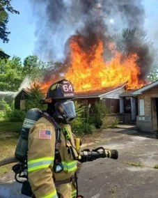 Firefighter in full gear standing in front of a house with a large fire and thick smoke in the background.