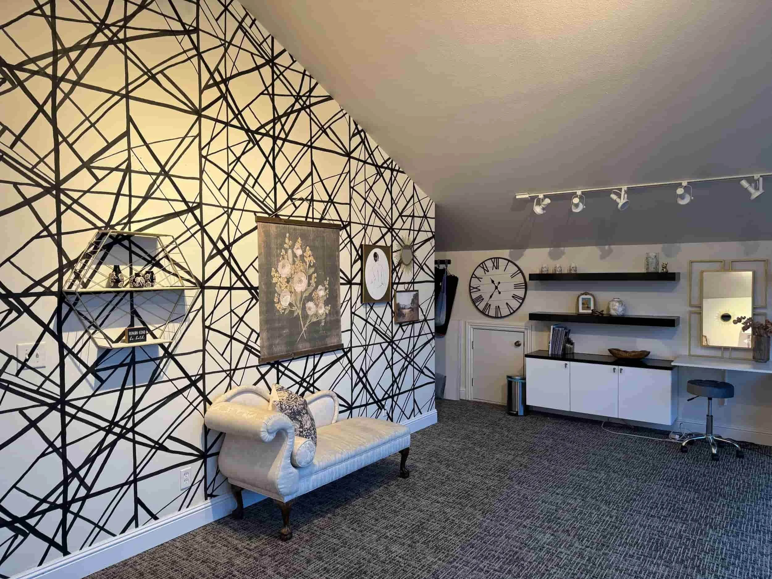 Living room with black geometric patterned wallpaper, white upholstered chaise lounge, framed artwork, wall clock, black shelving, and various decor items.