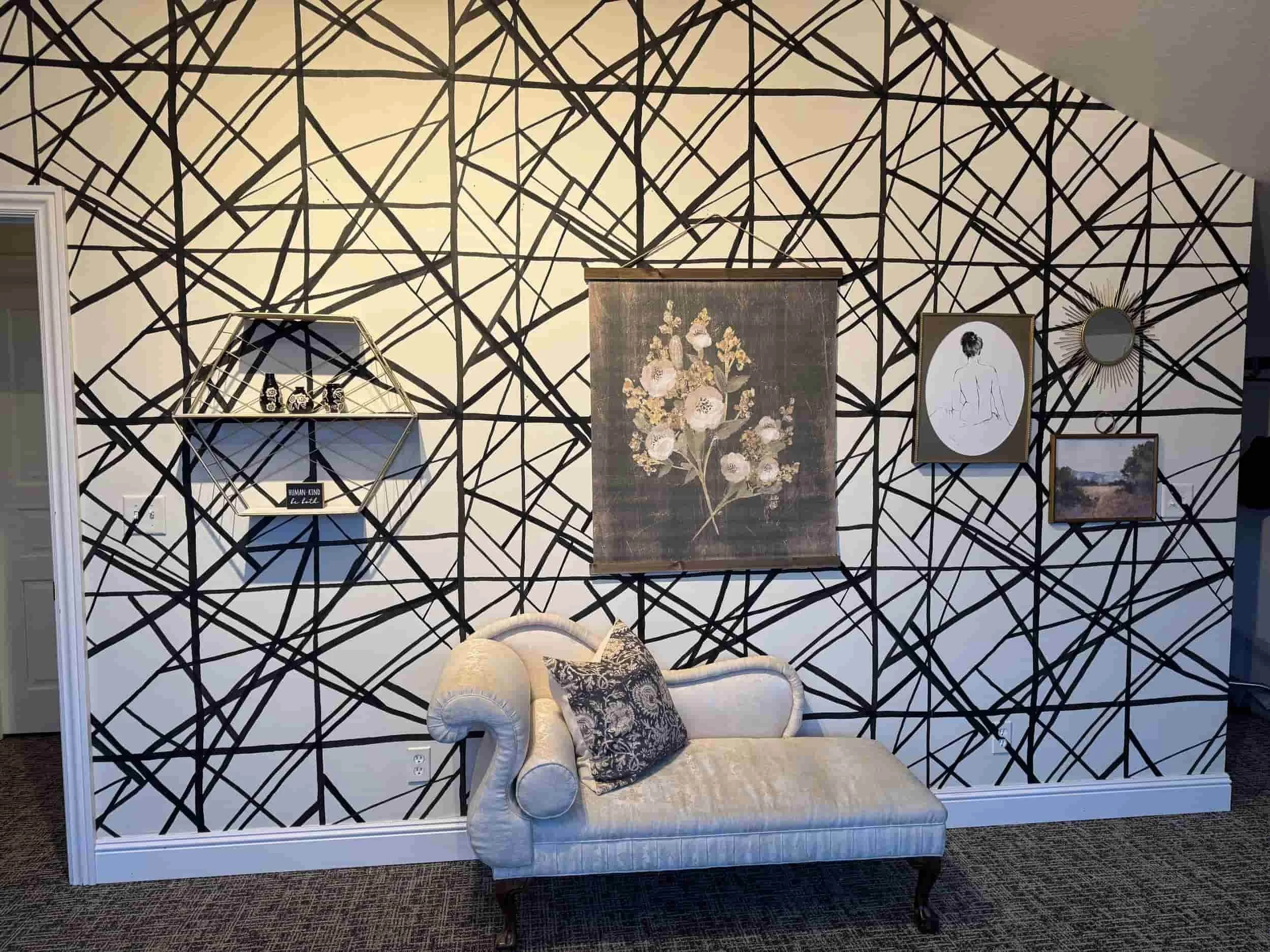 A living room wall with a black abstract line pattern on a white background, decorated with various framed artworks including floral art, a line drawing of a woman, a mirror, and landscape photos, with a white armchair and decorative pillows beneath.