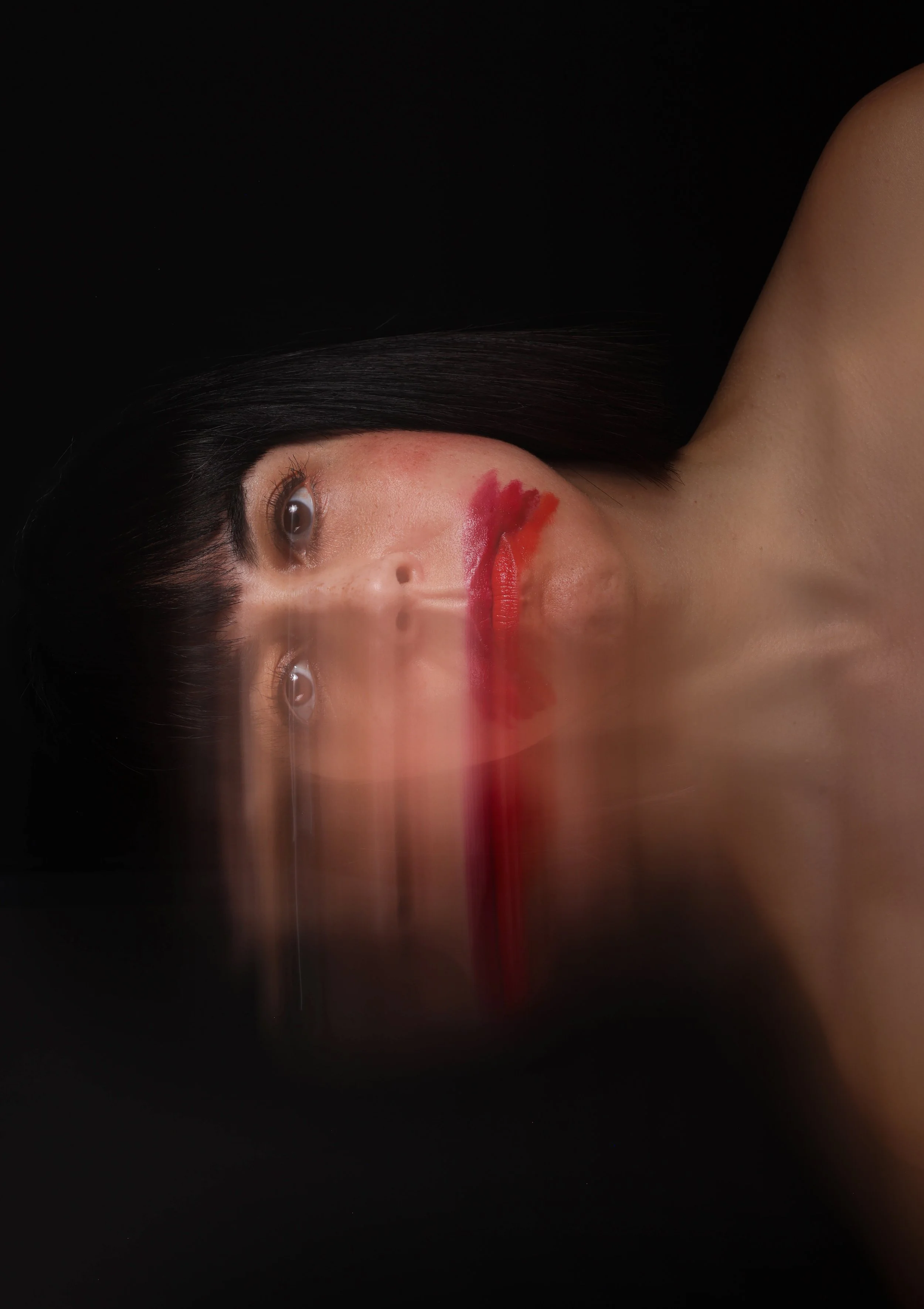 A woman with black hair, her face resting on a reflective black surface, with red makeup smeared across her lips and chin, and a reflection creating a symmetrical effect.