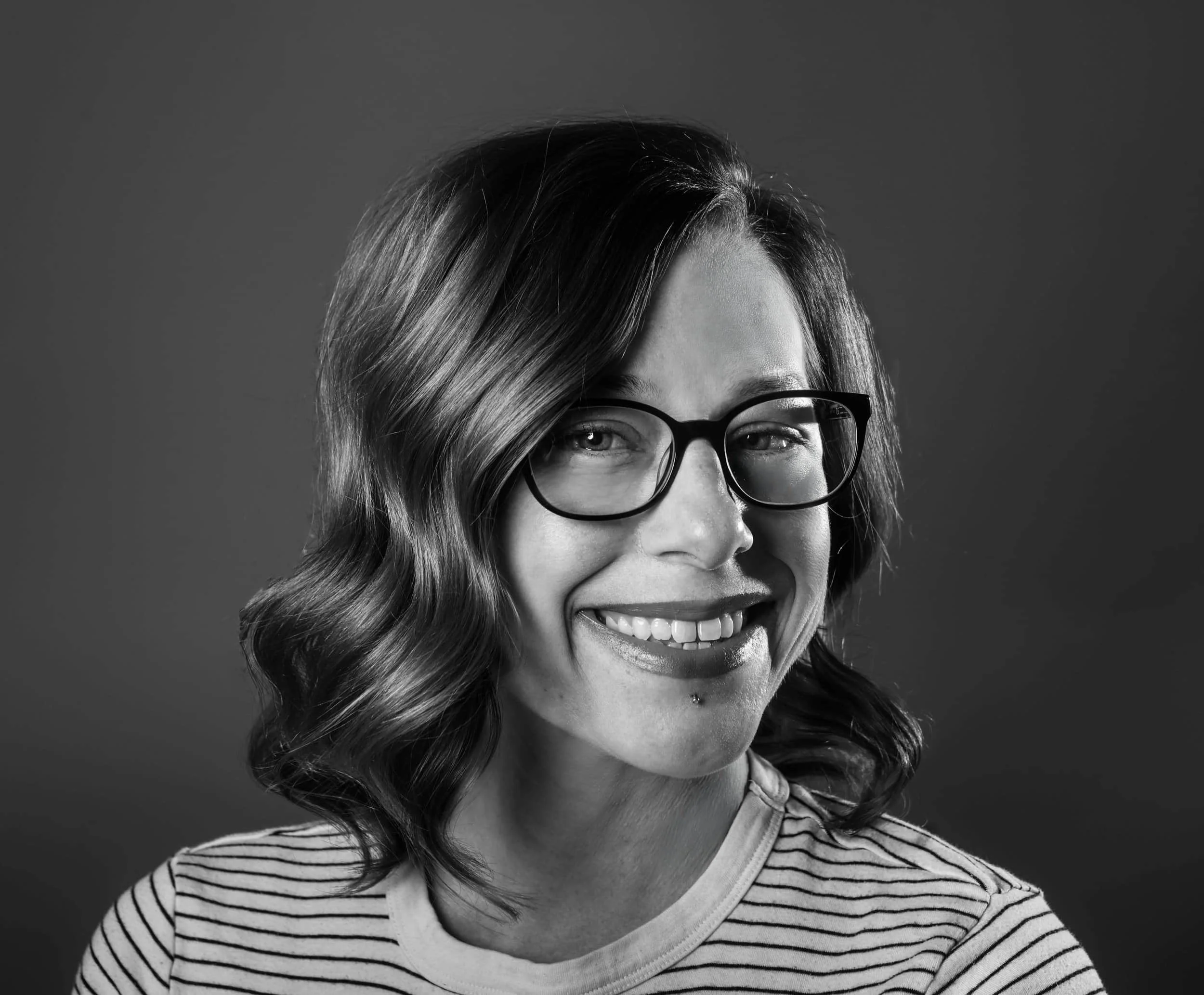 Black and white photograph of a smiling woman with glasses and wavy hair, wearing a striped shirt.