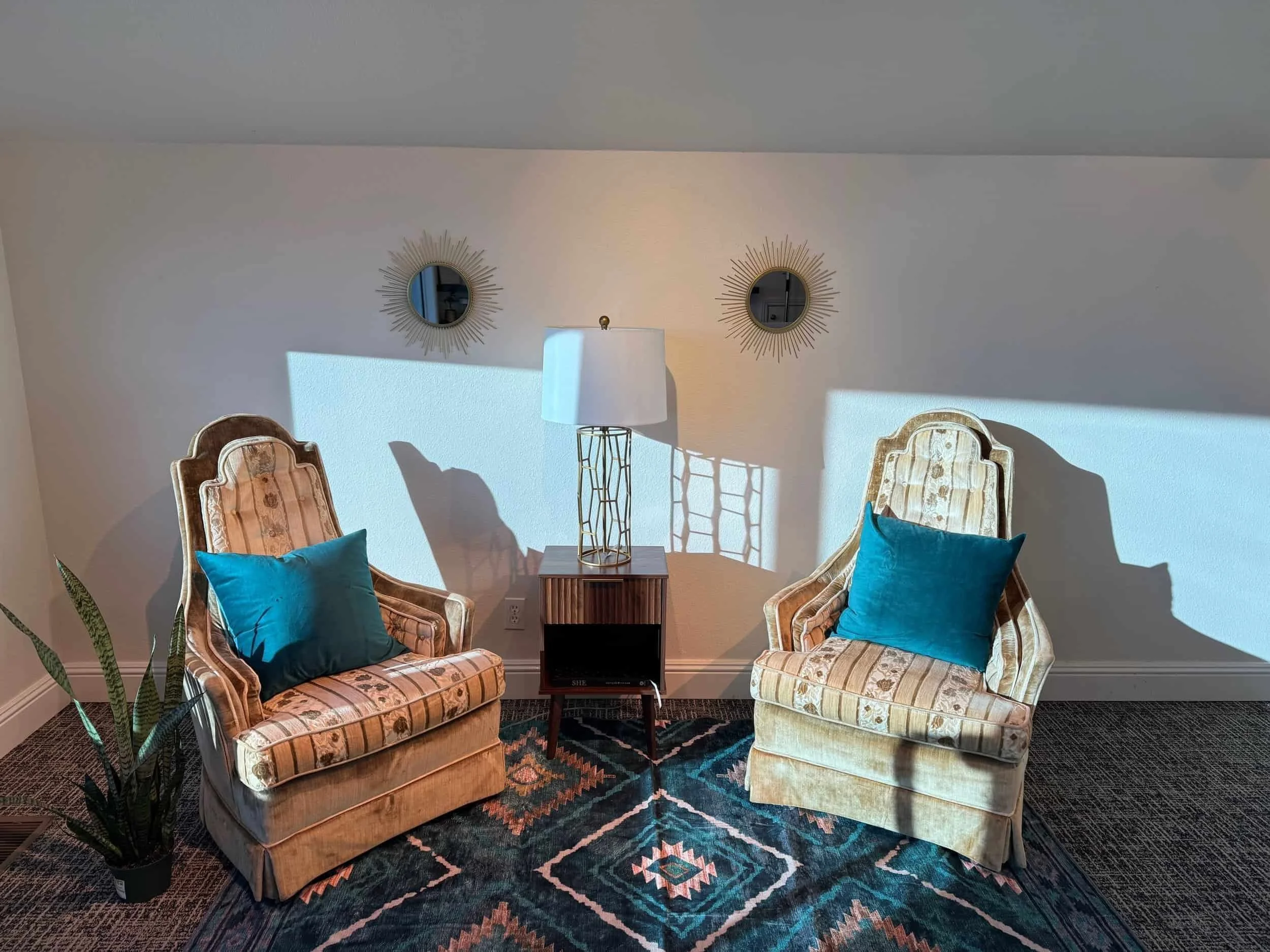 A living room corner with two vintage patterned armchairs having teal pillows, a small wooden side table, a white standing lamp, two decorative round mirrors with sunburst frames on the wall, a rug with a geometric pattern, and a plant in the corner.
