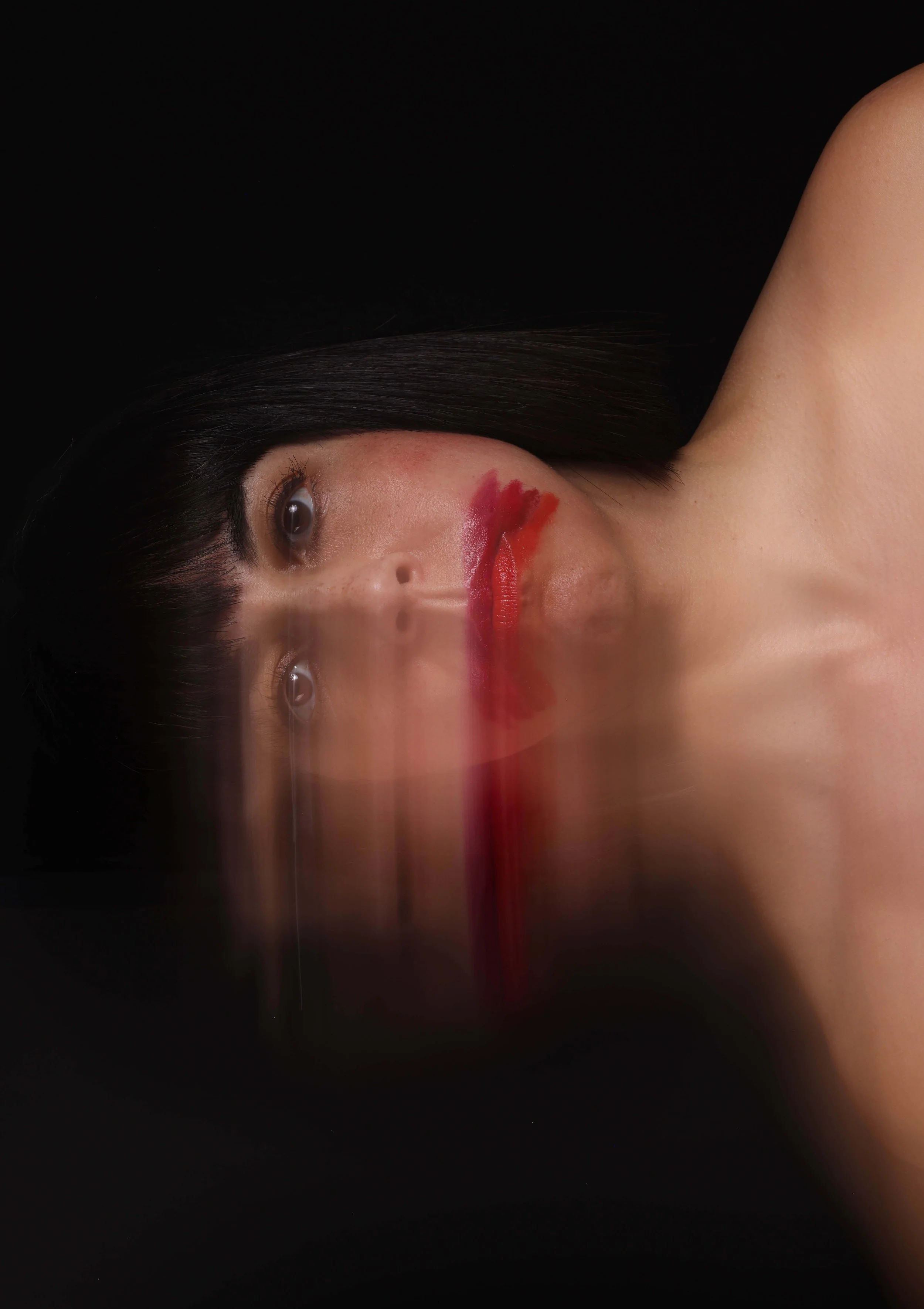 A woman with black hair and light skin lying on her side against a black background, with red paint or makeup smudged across her face and neck.