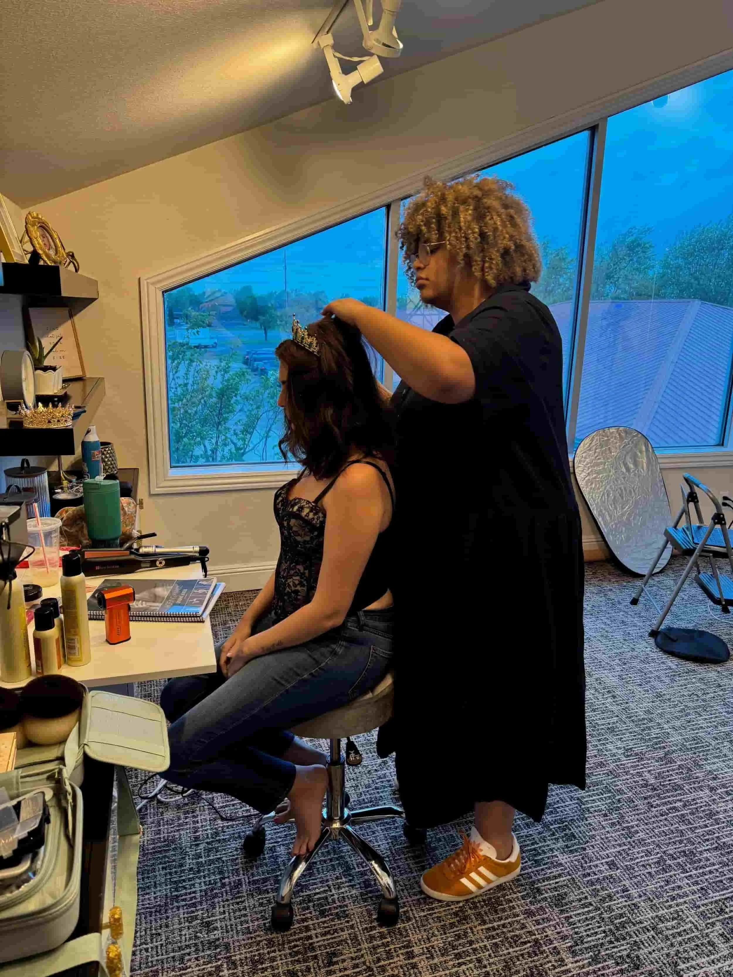 A woman is seated while another woman styles her hair with a tiara. The seated woman is dressed in a black lace top and jeans, with her hair partly styled and a tiara on her head. They are in a room with large windows and various hair and beauty supplies on a table nearby.