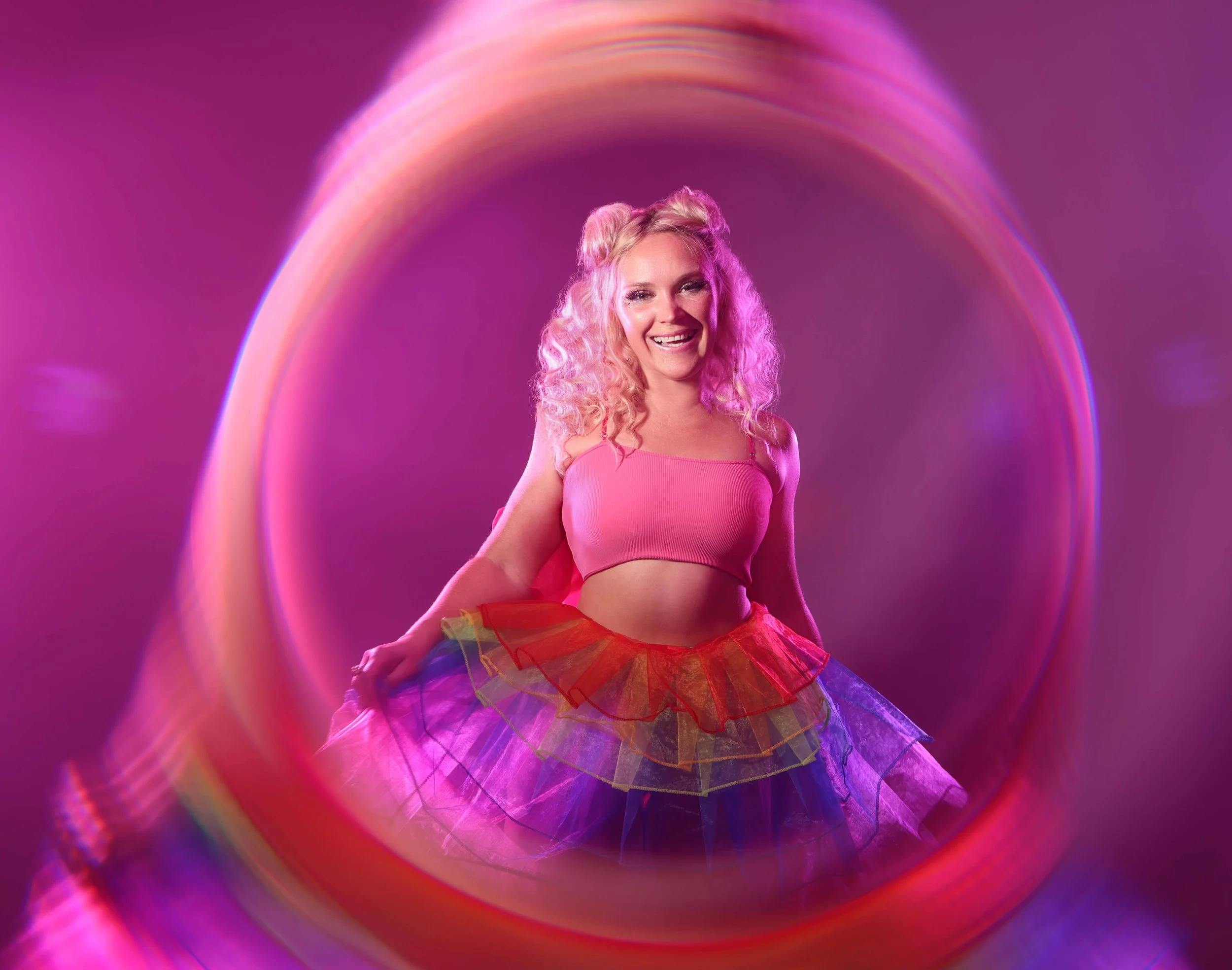 A woman with pink hair wearing a pink top and a rainbow-colored tulle skirt, standing in front of a purple background with a rainbow-colored light effect surrounding her.