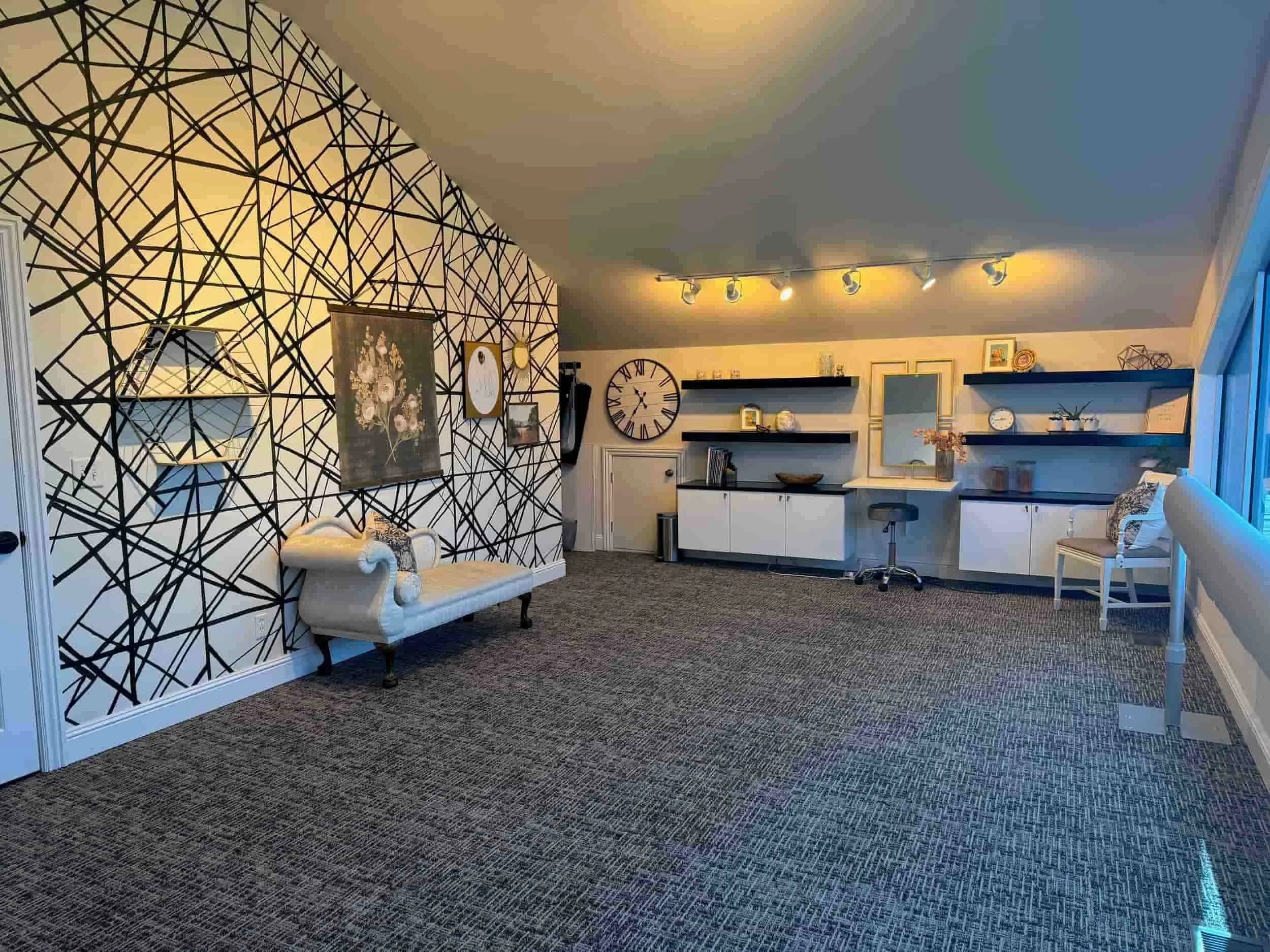 Living room with geometric black and white accent wall, beige sofa with decorative pillows, black wall clock, and floating shelves with decor items, illuminated by track lighting.