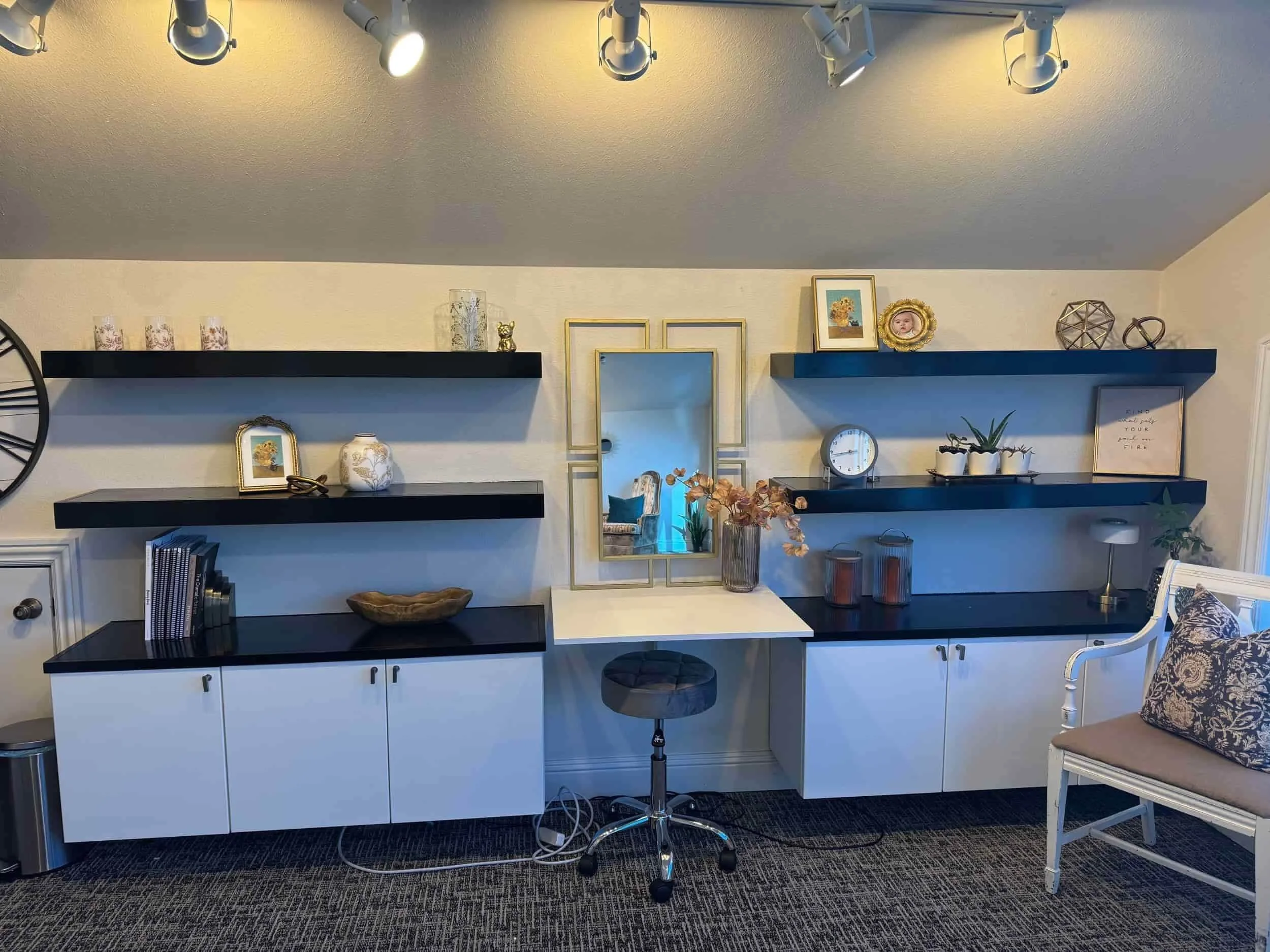 Living room with beige walls and dark blue floating shelves decorated with framed pictures, vases, and various ornaments. A black and white clock is on the right shelf, and a large mirror with a gold frame is in the center. There is a white cabinet b