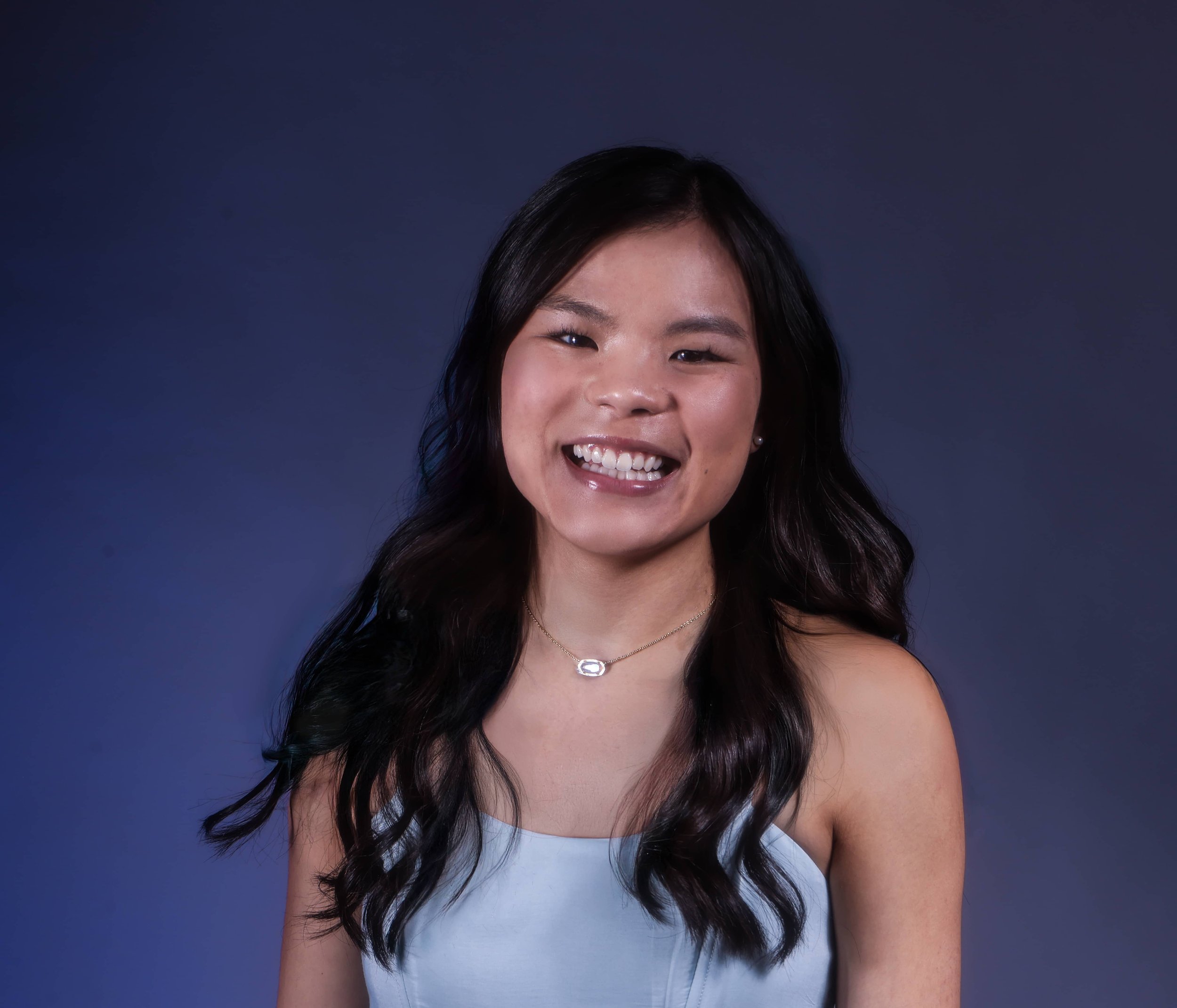 Close-up of a smiling young woman with long dark hair, wearing a light blue top and a silver necklace, against a dark blue background.