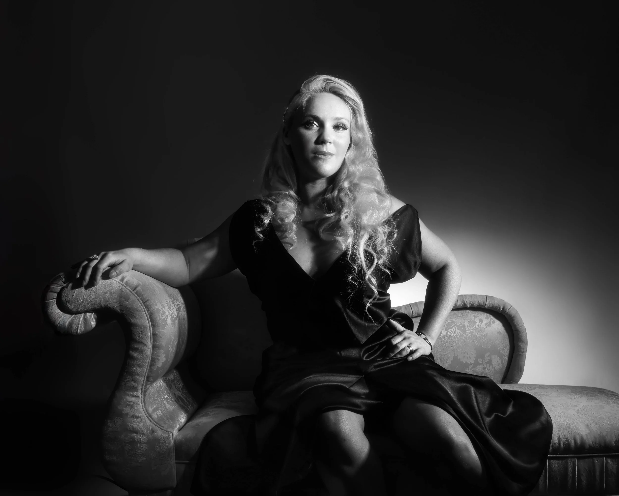 Black and white portrait of a woman with long curly blond hair sitting on a chaise lounge 