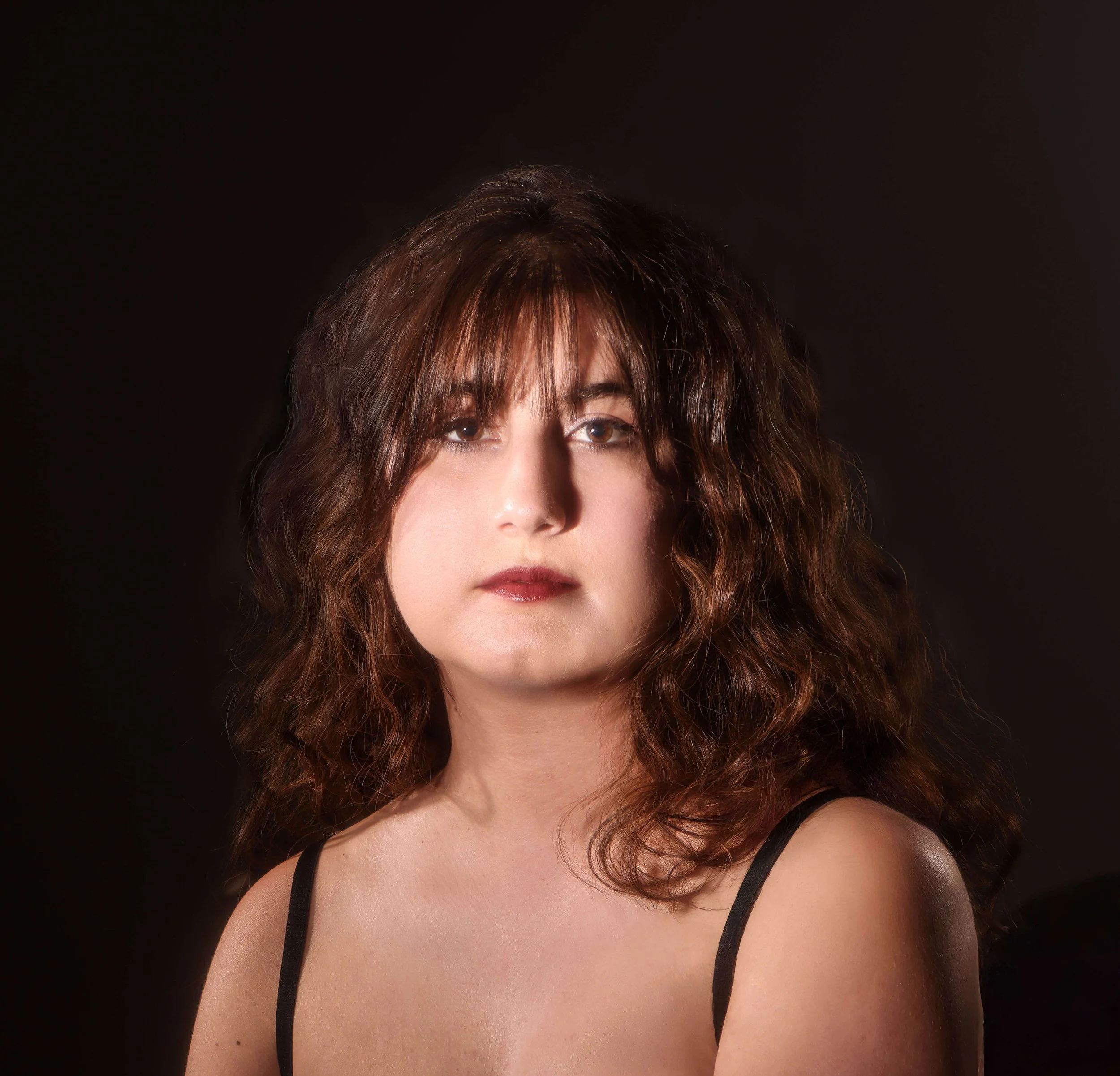 Close-up of a woman with curly brown hair wearing a black tank top against a dark background.