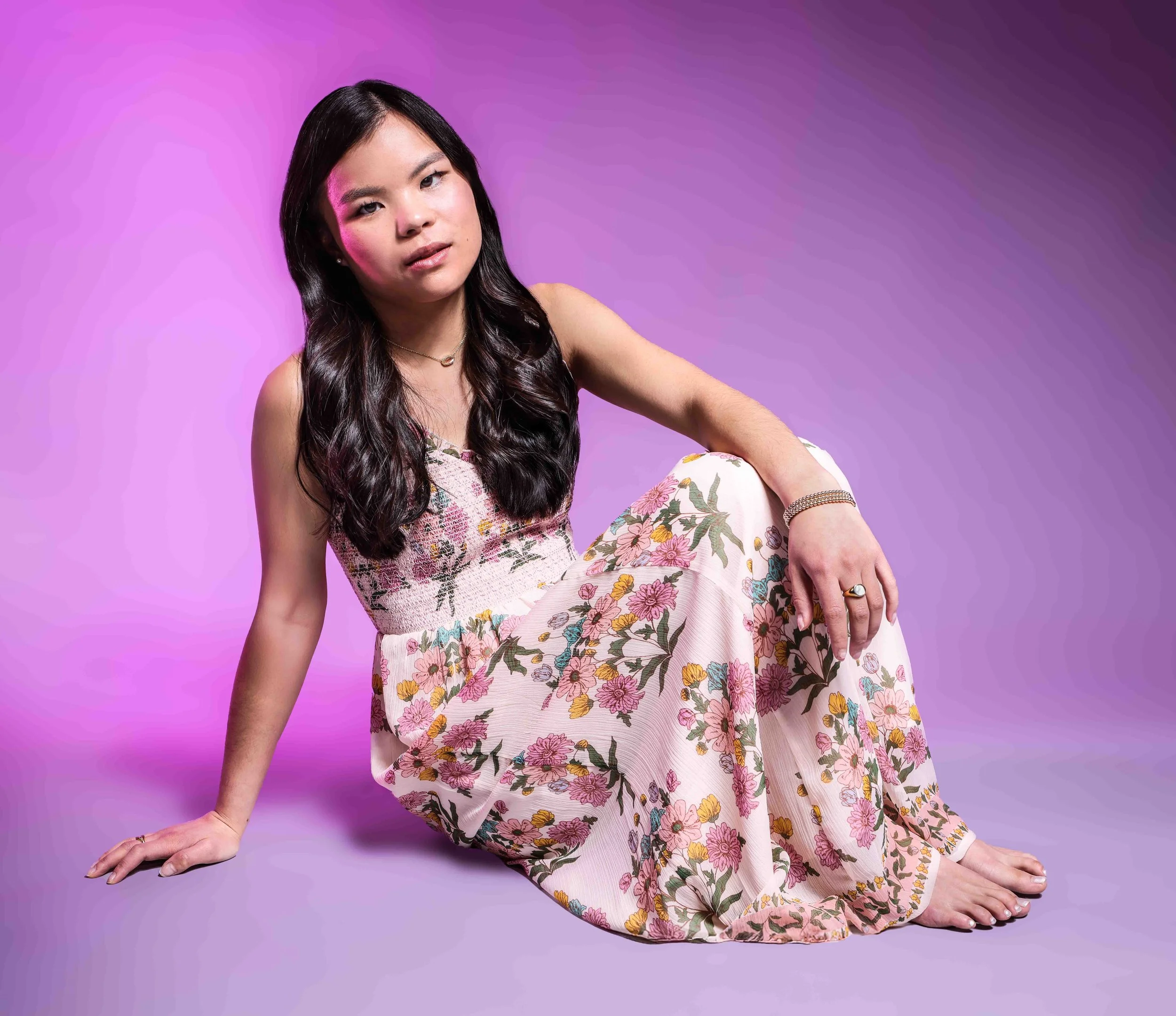 Portrait of an Asian American girl sitting wearing a floral dress on a pink background. 