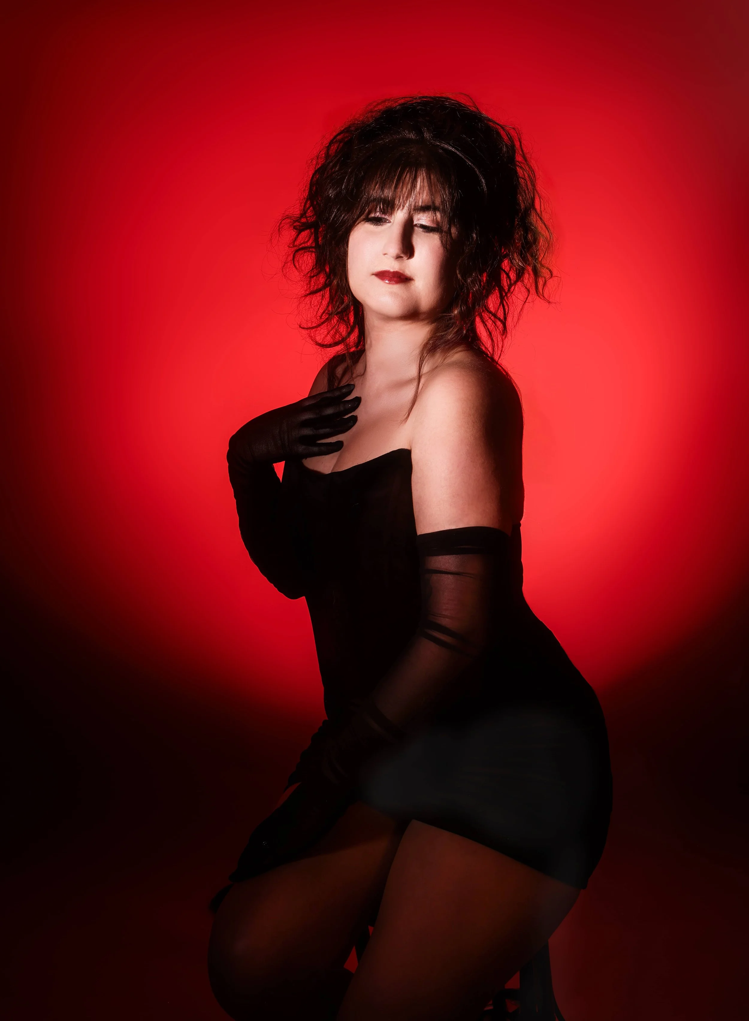 A woman with curly dark hair, wearing black gloves and a black dress, posed against a red background with a seductive expression.