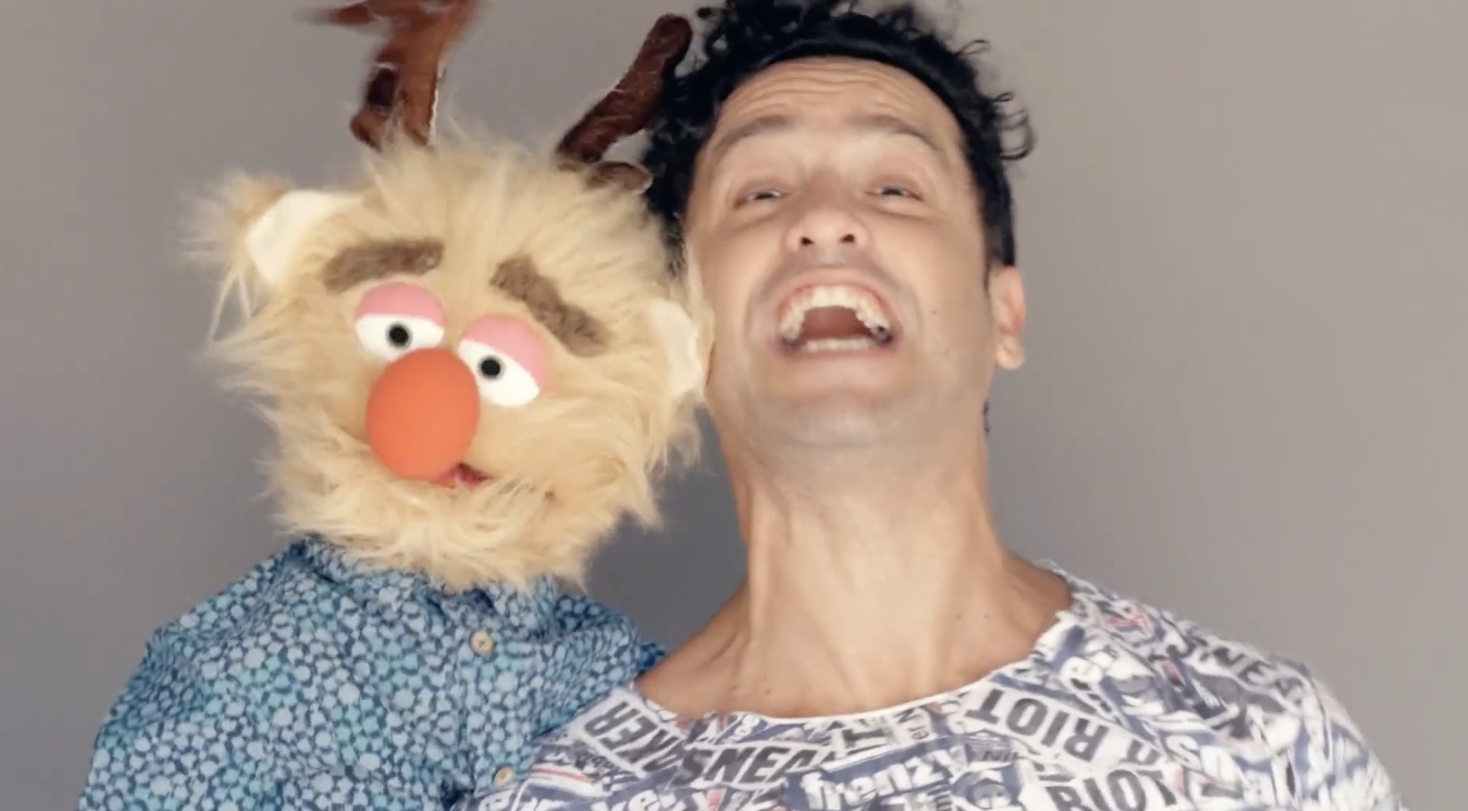 A man with curly black hair and a woman with a puppet face pose together, smiling and laughing.