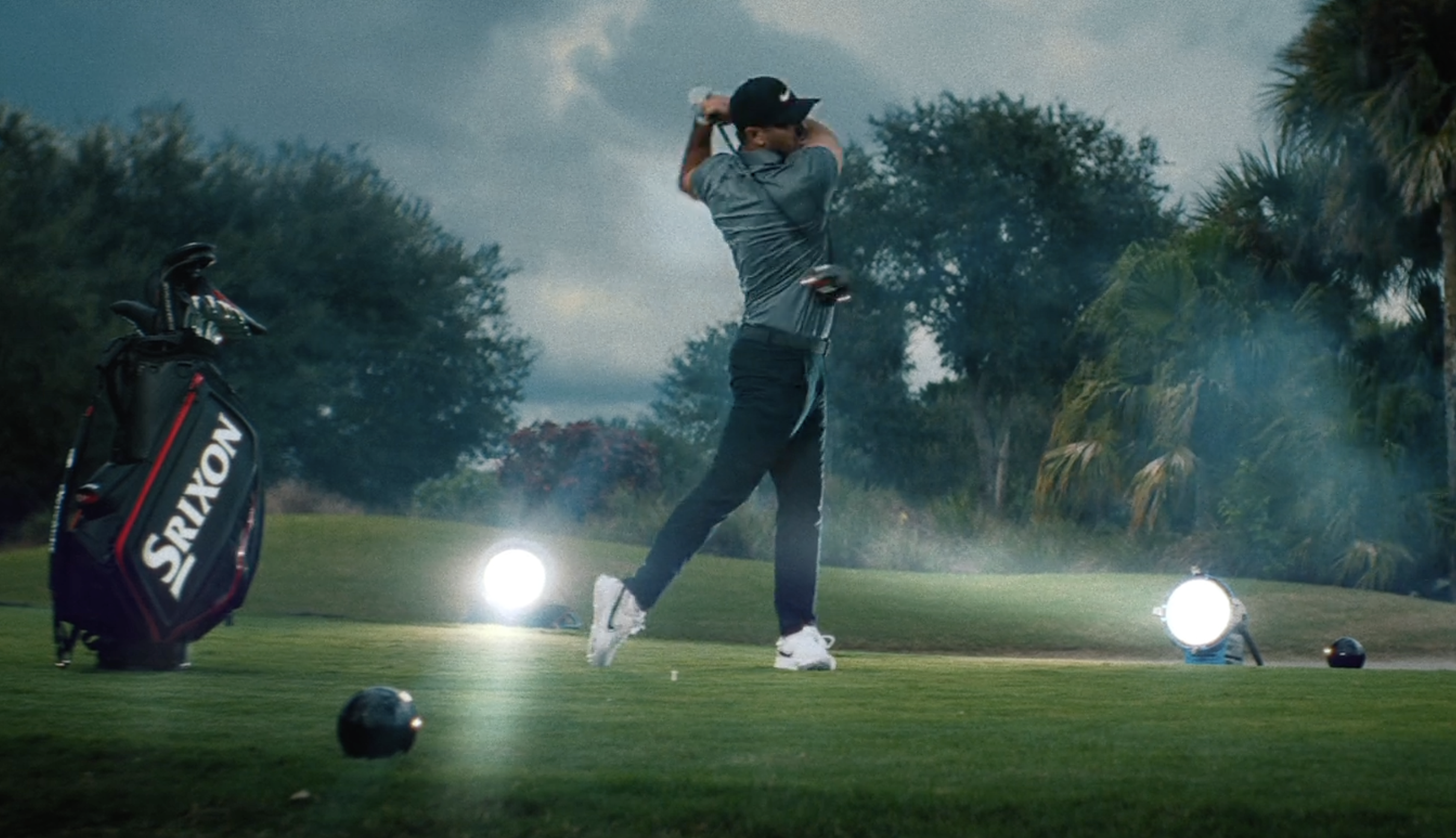 A man in a gray shirt and black cap swinging a golf club on a golf course during dusk, with a golf bag and bright lights in the background.